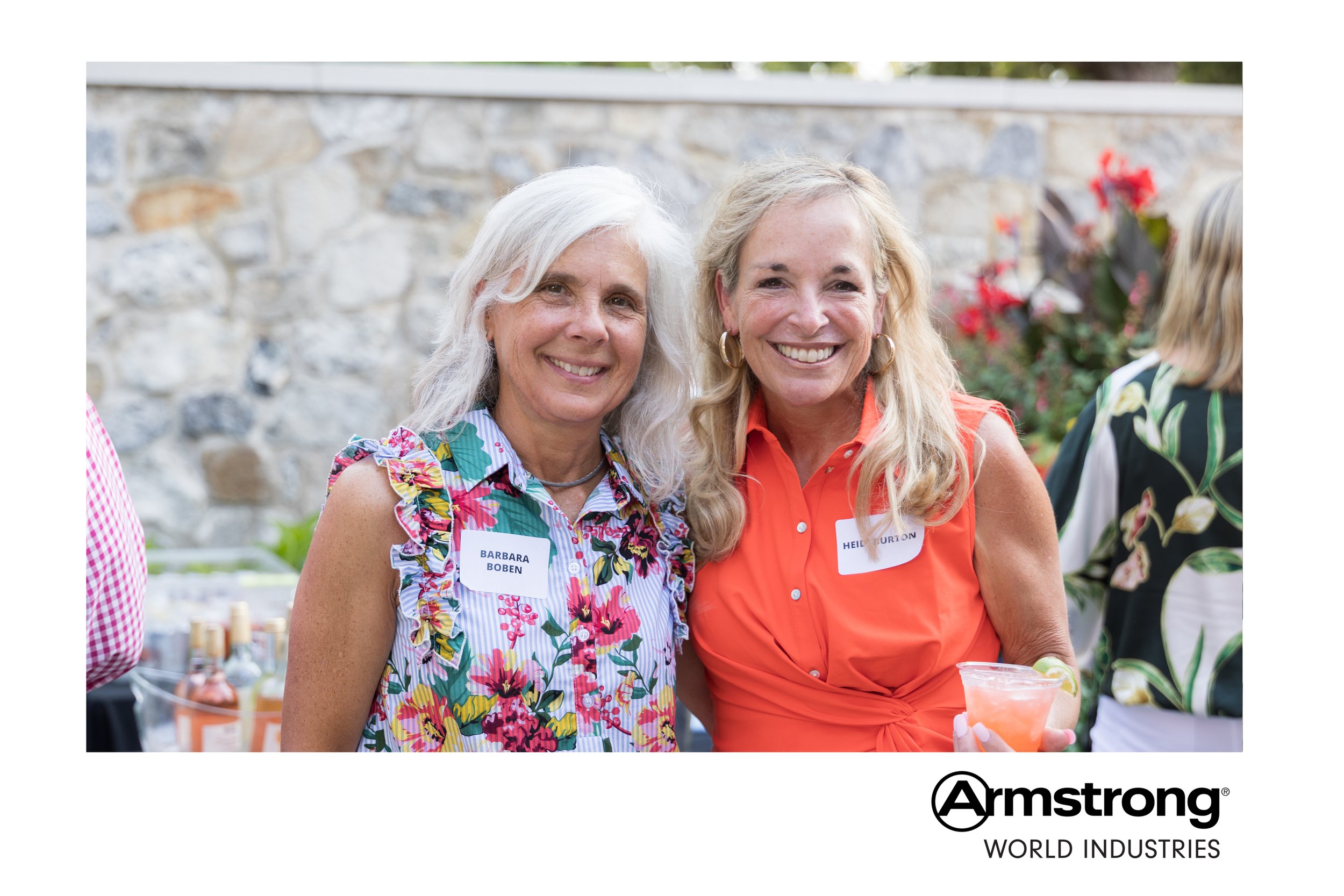 Two smiling women with name tags, standing outdoors at a social event, with flowers and a stone wall in the background, one holding a pink cocktail.