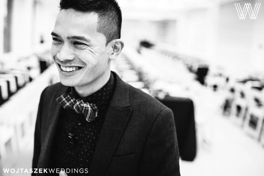 A smiling man wearing a dark suit, patterned shirt, and bow tie, in an office or conference room setting.