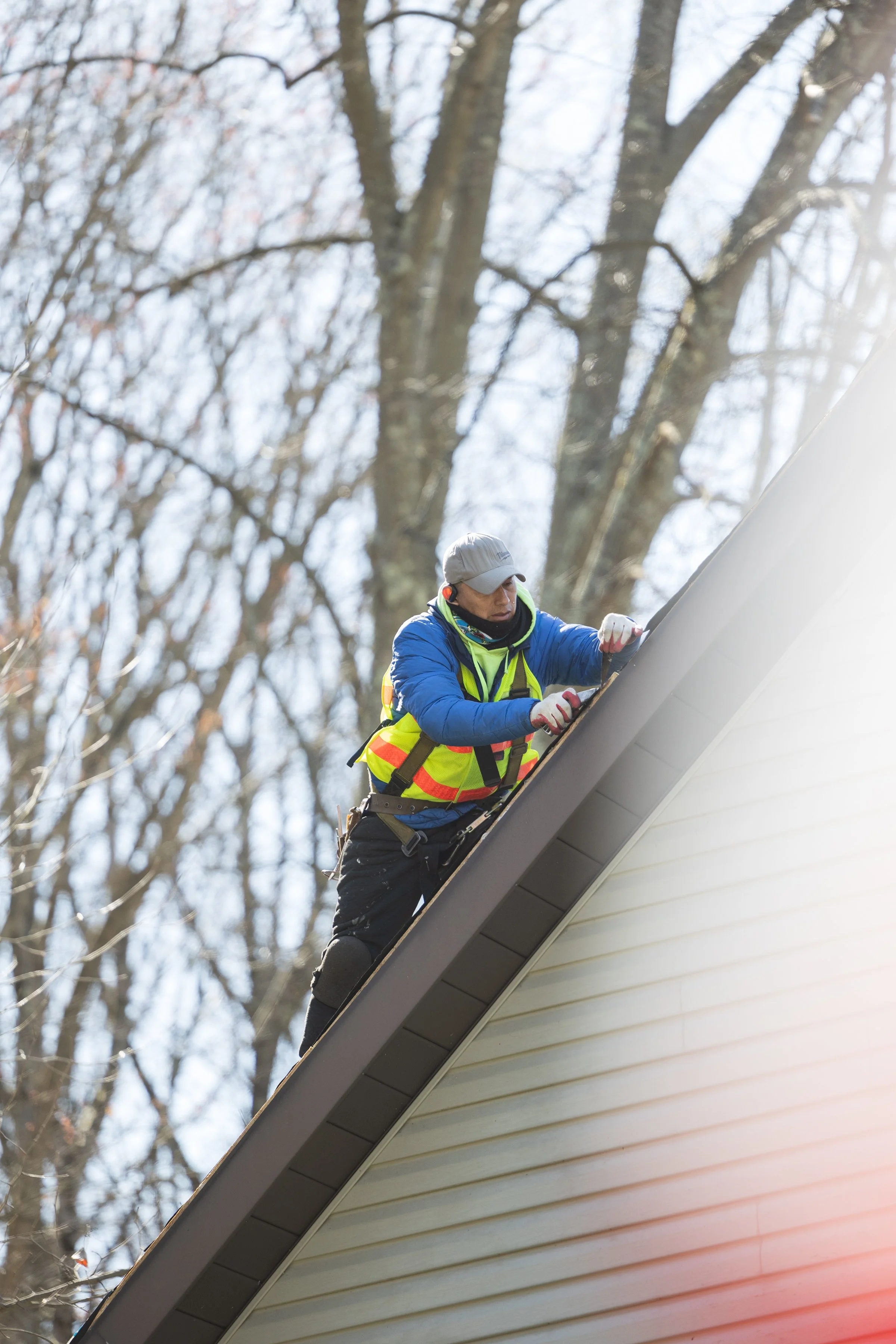 A worker wearing a gray cap, a blue jacket, and a high-visibility safety vest is repairing or inspecting the roof of a house. The worker is standing on the sloped roof, focused on the task, with a background of tall, leafless trees and a cloudy sky.