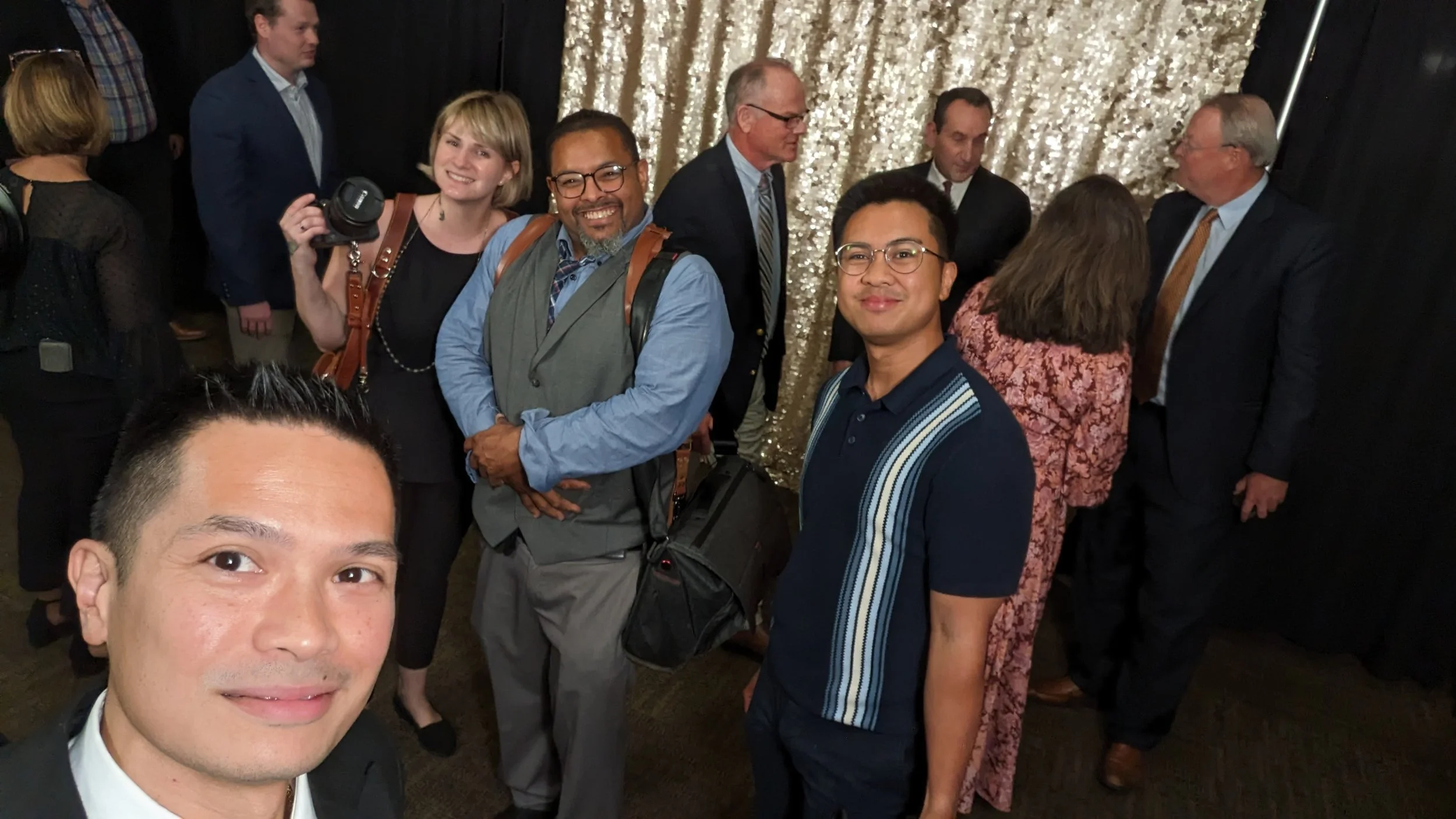 Group of people at an indoor event with a sequin backdrop, including three smiling men in the foreground and others talking in the background.