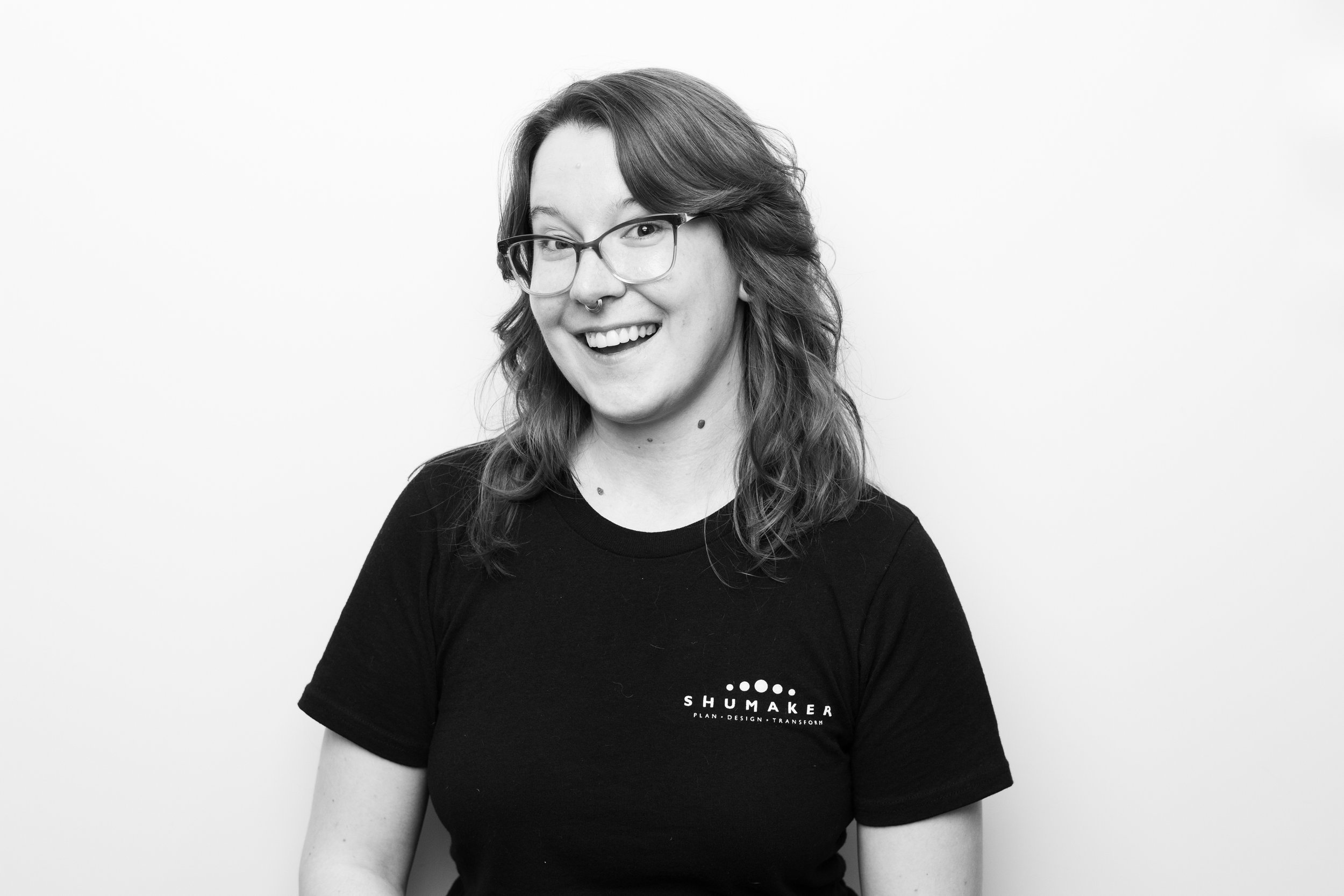 Black and white photo of a woman with glasses, smiling, wearing a black T-shirt with the words "SHUMAKER" and a slogan underneath, in front of a plain background.