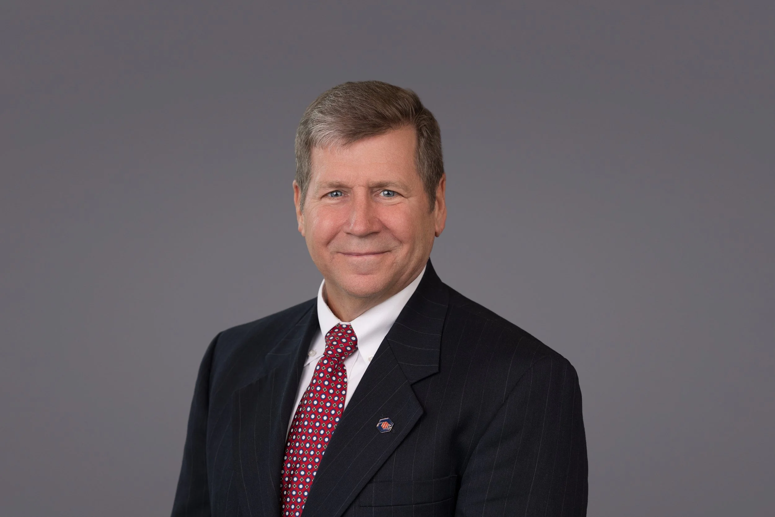 A professional headshot of a man in a suit with a dark pinstripe jacket, white shirt, and red patterned tie, smiling against a plain gray background.