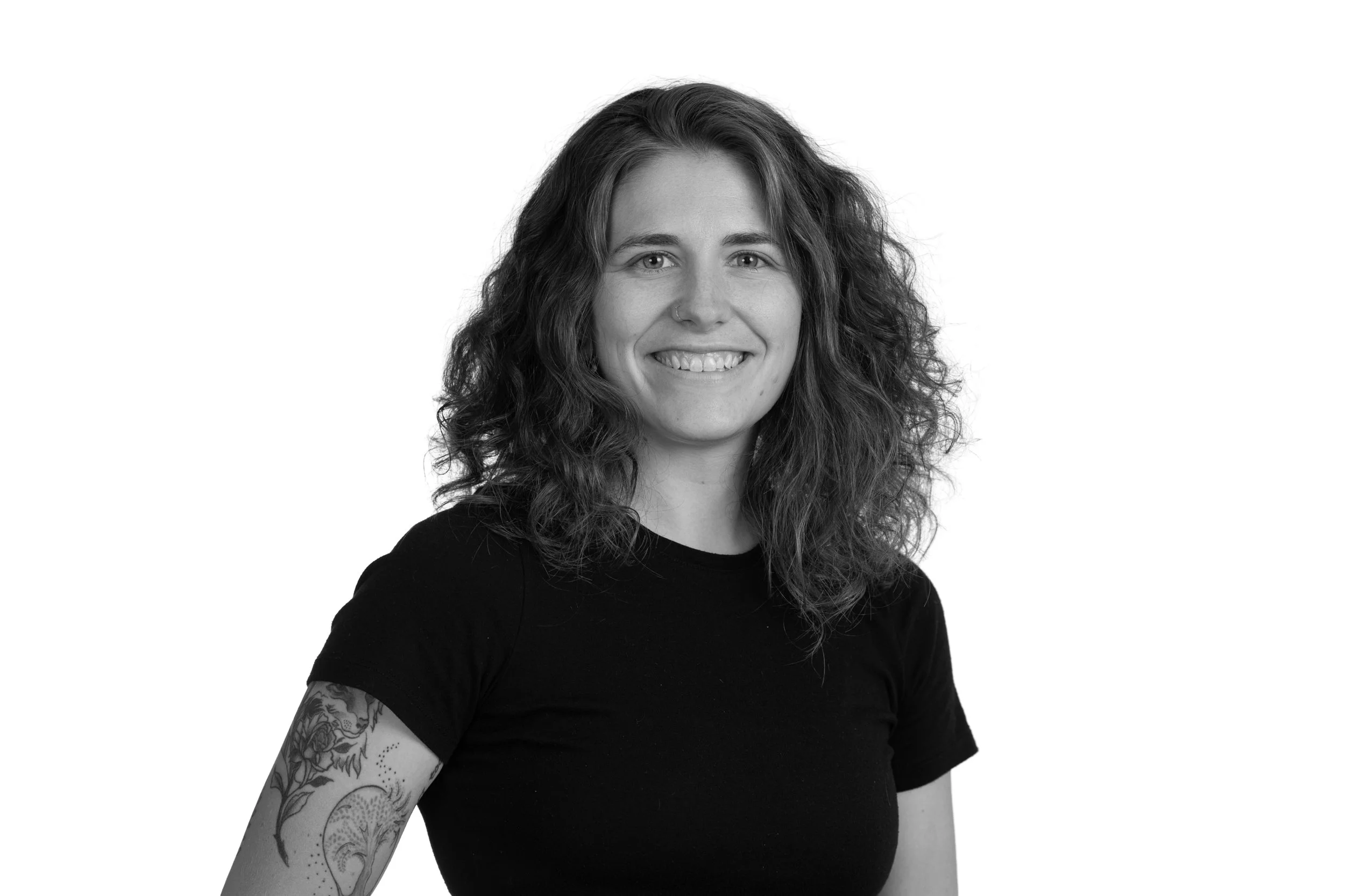 Black and white portrait of a young woman with curly hair, smiling, wearing a black t-shirt, with tattoos on her left arm, against a plain white background.