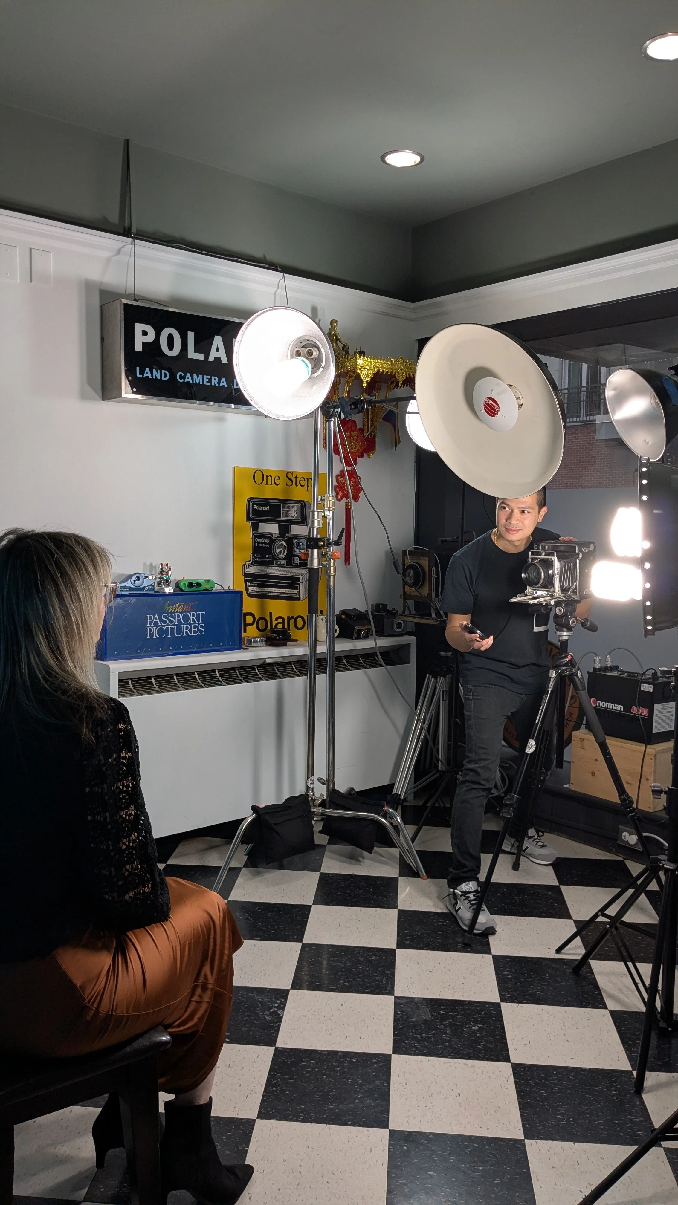 A woman sitting on a chair in a photography studio being photographed by a man with a camera. The studio has various lighting equipment, a Polaroid sign, and a checkered black-and-white floor.