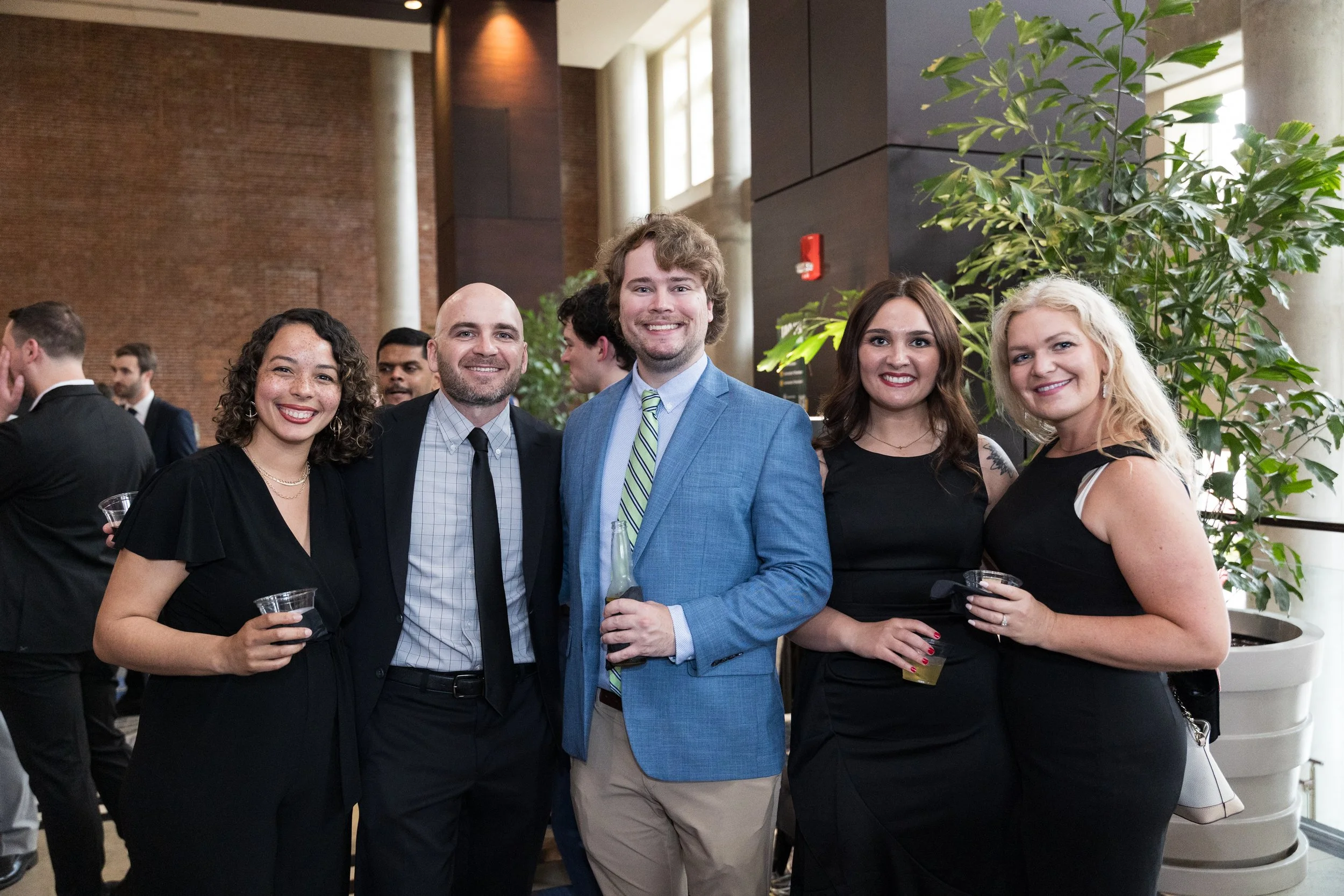 Group of five young adults dressed in formal attire at a social event, smiling, standing indoors near a large plant.
