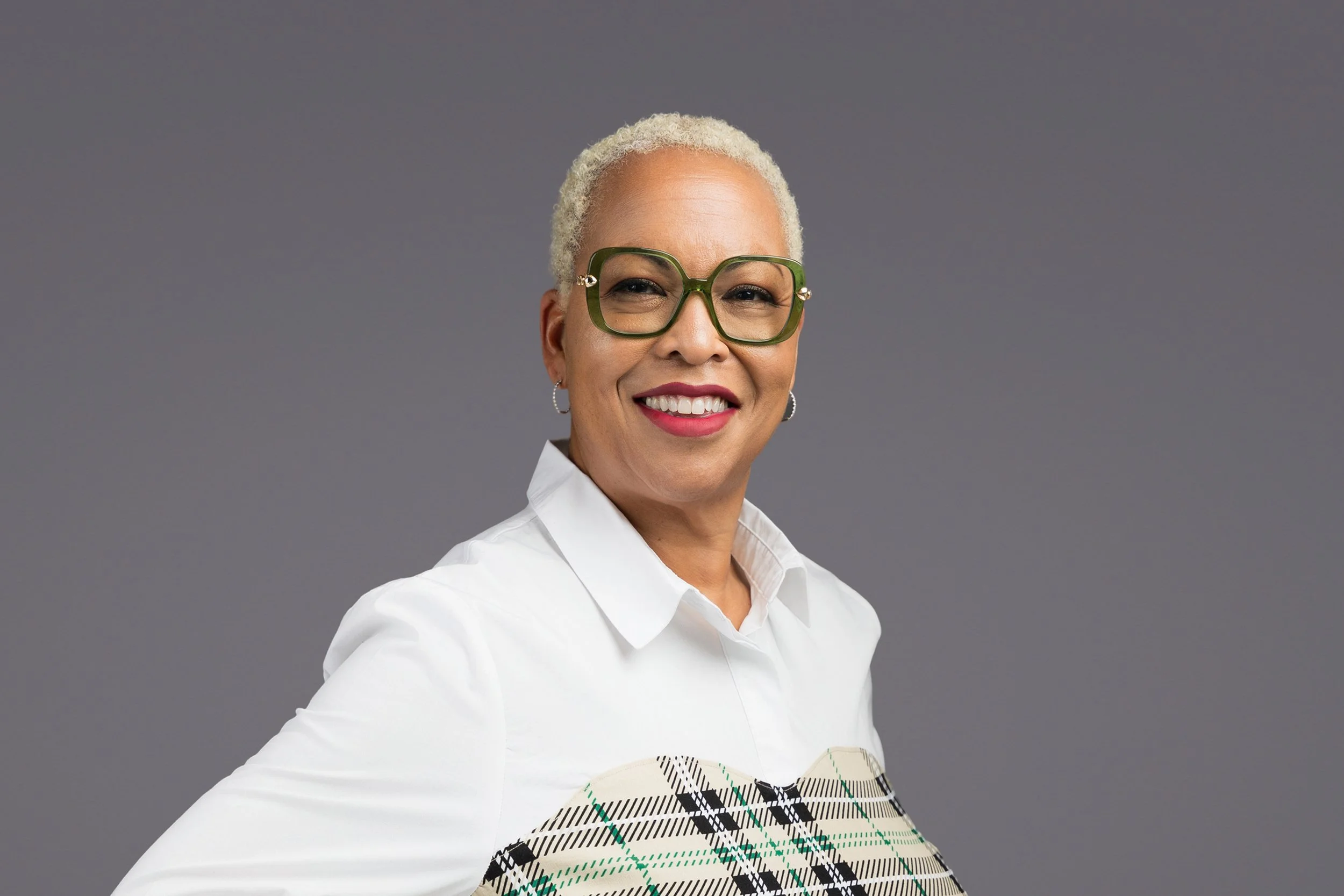 A smiling woman with short, curly, blonde hair wearing large green glasses, a white blouse, and a blazer with a plaid pattern, standing against a gray background.