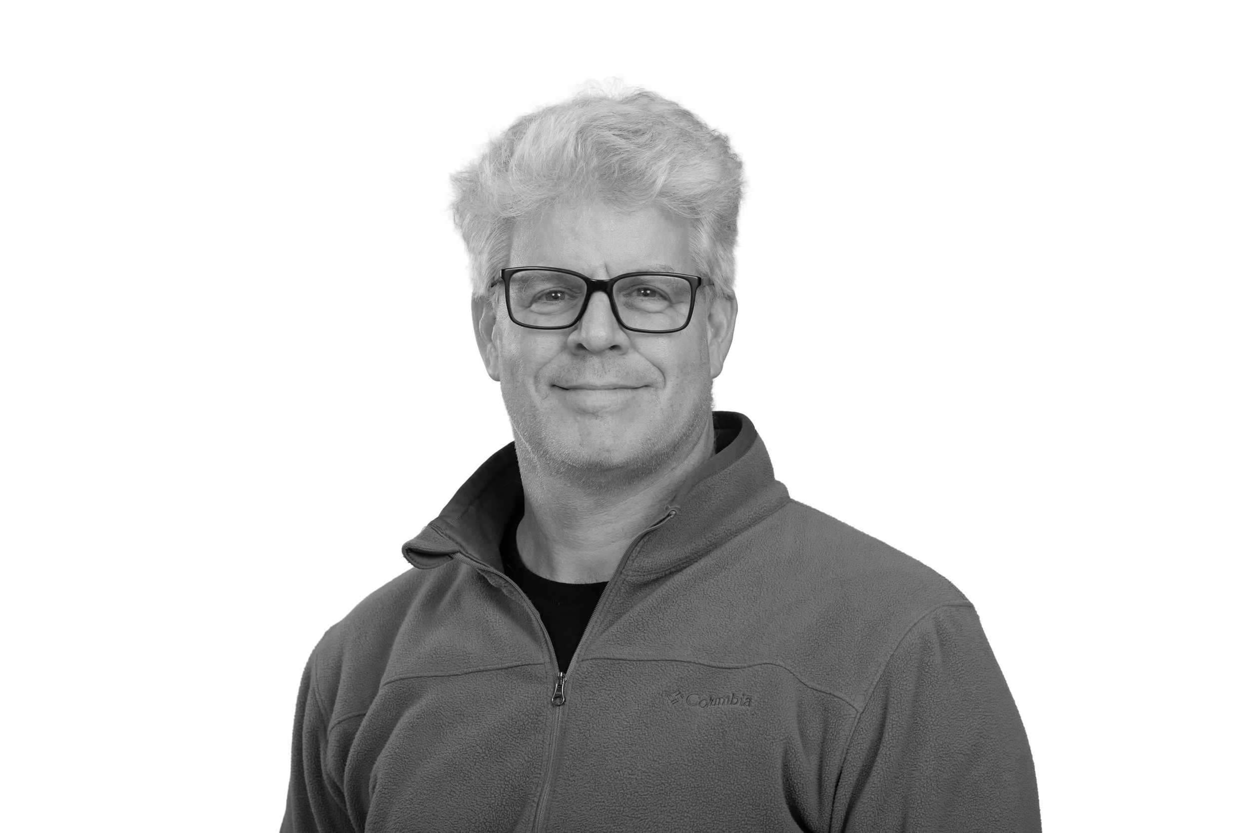 Black and white portrait of a middle-aged man with gray hair, wearing glasses and a Columbia fleece jacket, smiling at the camera against a plain white background.
