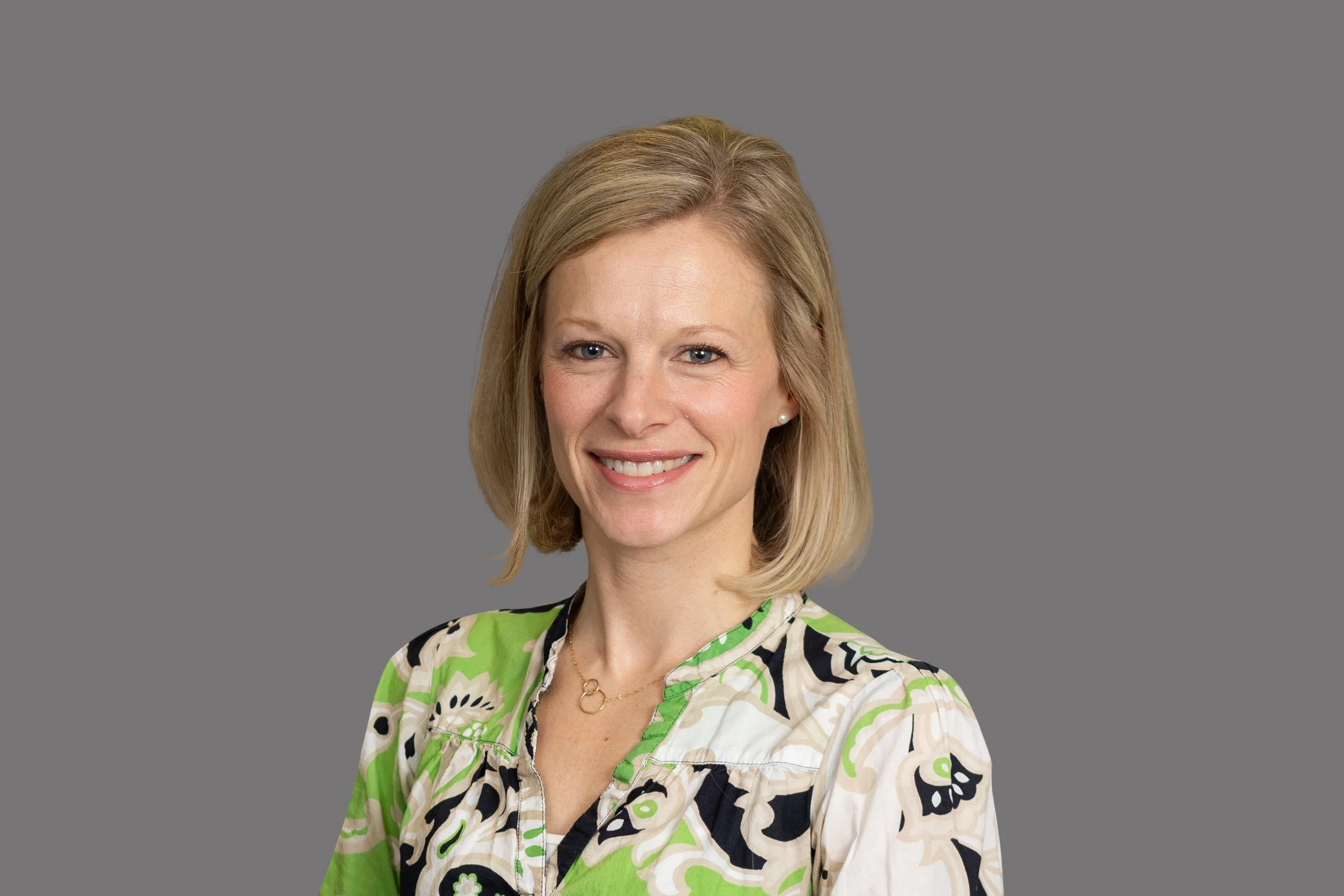 A woman with blonde hair smiling at the camera, wearing a colorful floral blouse with green, black, and white patterns, against a plain gray background.