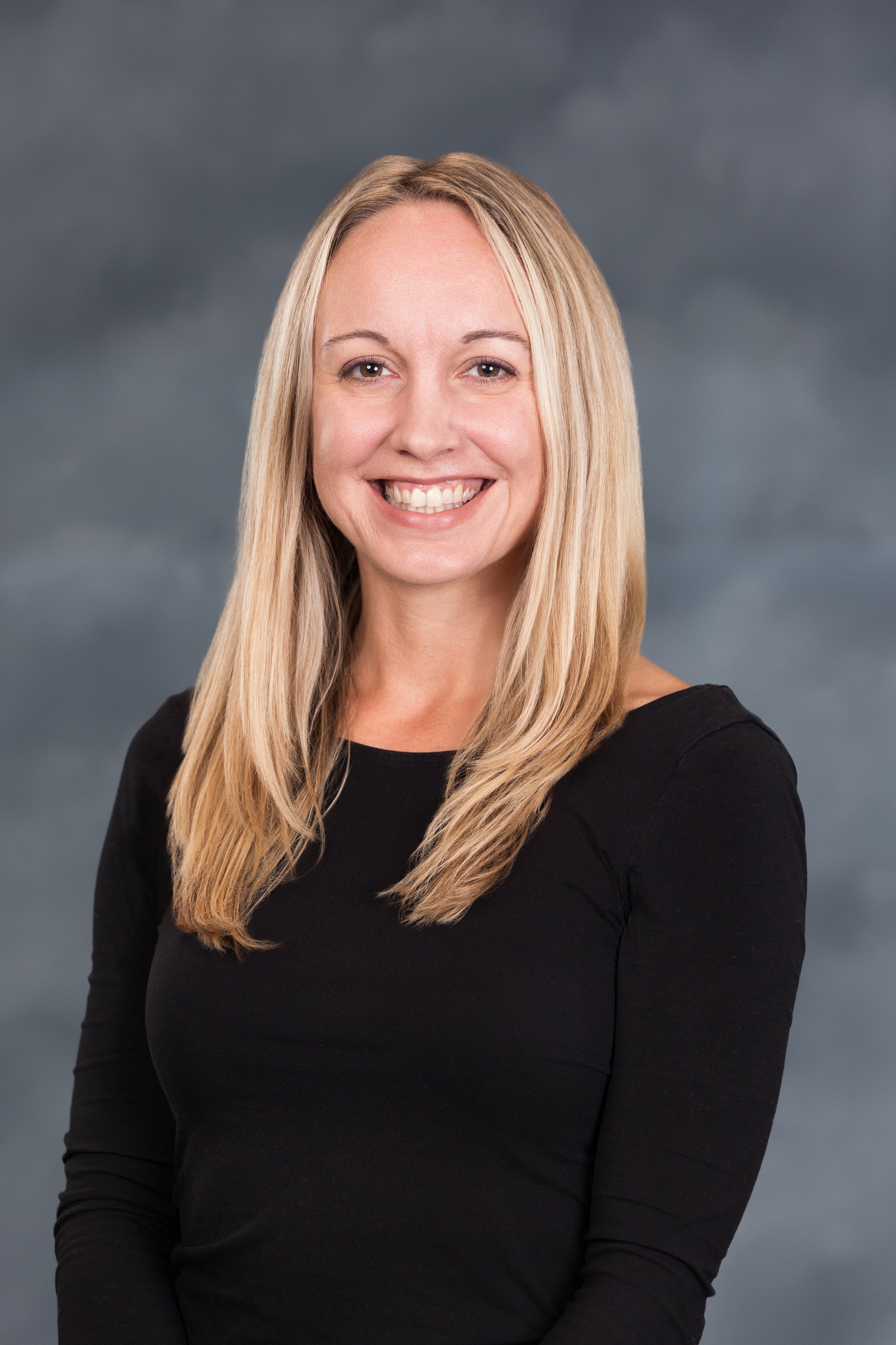 Headshot of a smiling woman with long blond hair, wearing a black top, against a dark cloudy background.