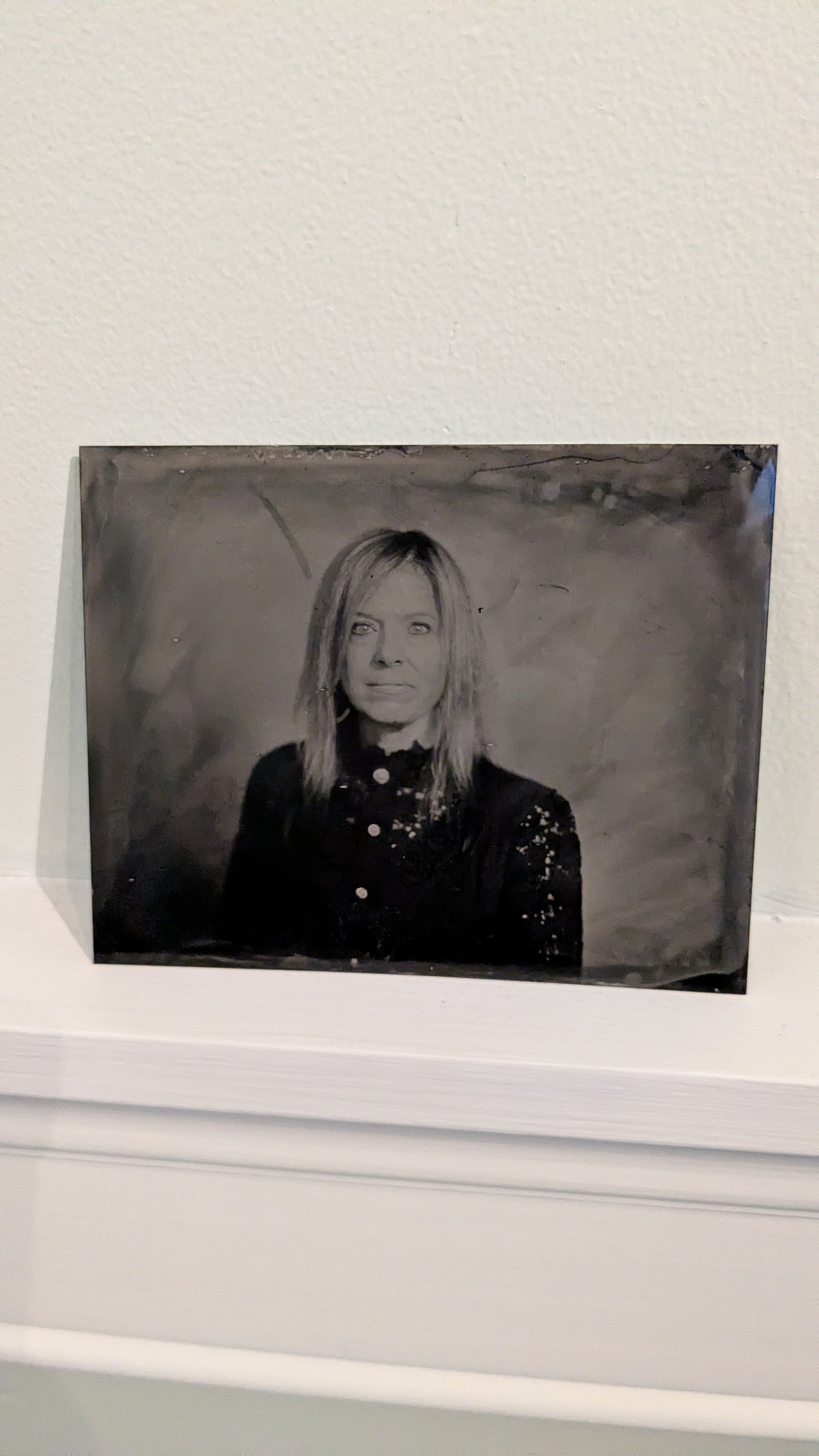 A black and white portrait photograph of a woman with shoulder-length hair, wearing a dark, possibly lace, top, placed on a white surface against a plain wall.