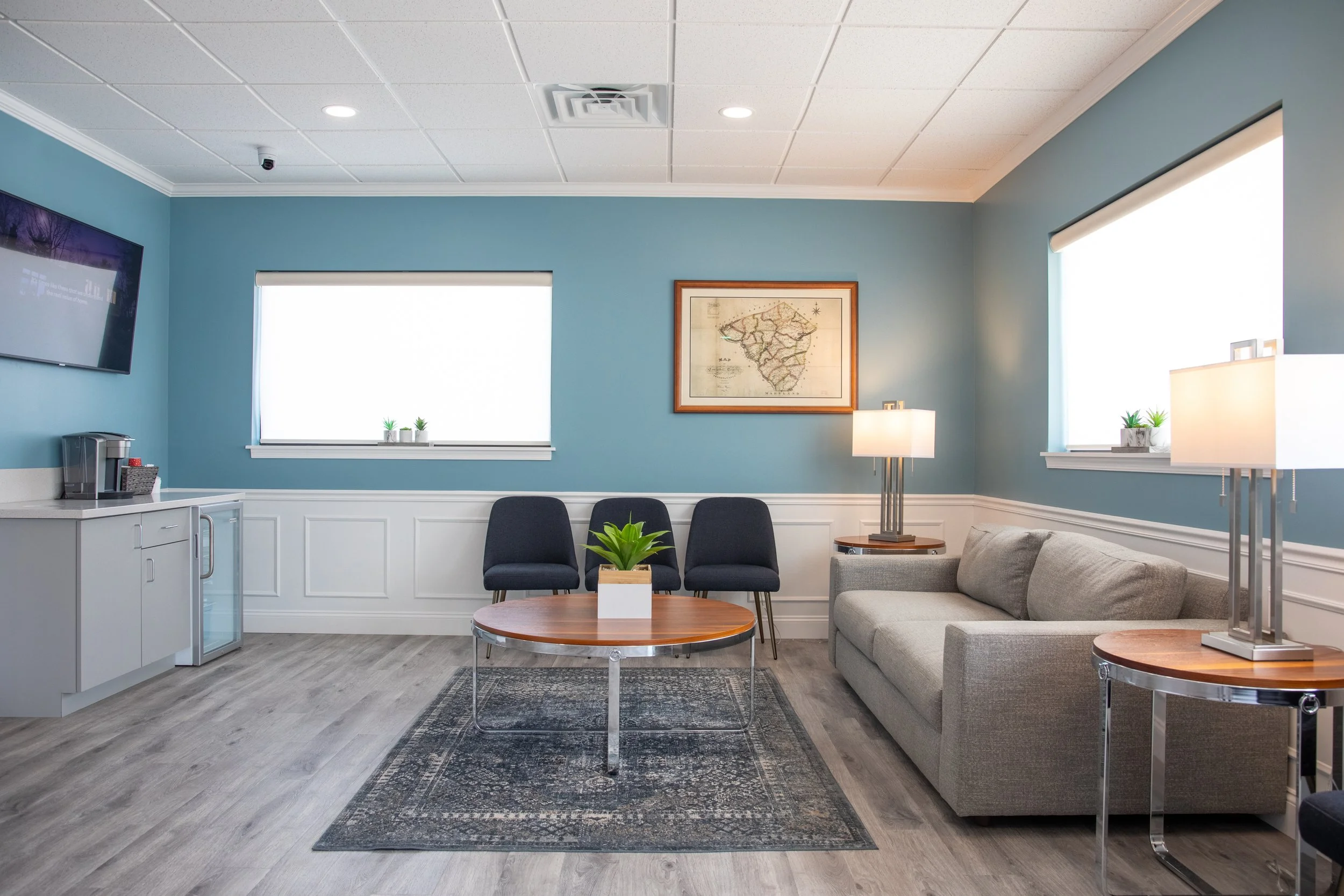 A waiting room with a gray sofa, three black chairs, a round wooden coffee table with a plant, a patterned rug, a lamp, a framed map on a blue wall, two windows with blinds, a wall-mounted TV, and a small kitchenette area with a coffee machine.