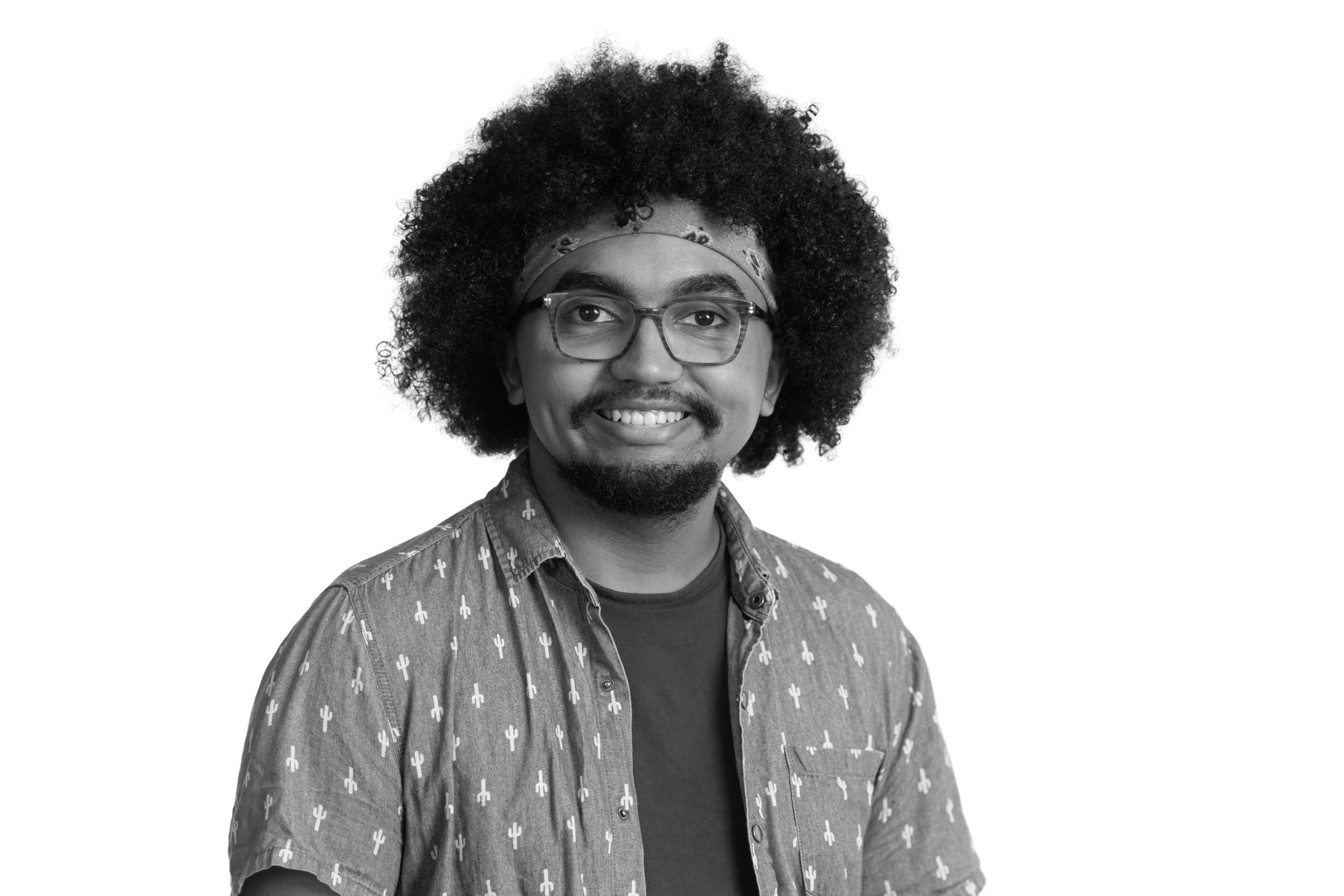 Black and white portrait of a man with curly hair, glasses, mustache, and goatee, smiling, wearing a patterned shirt and a bandana headband, against a plain white background.