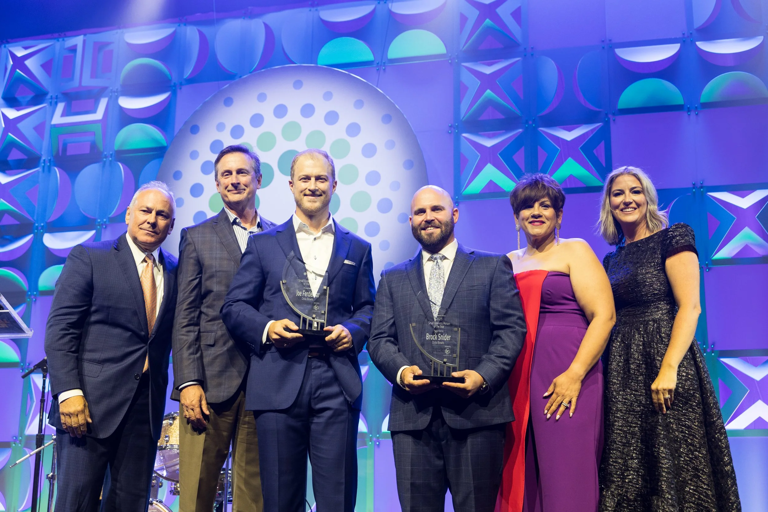 Group of six people on stage at an award ceremony, with two men holding awards, posing for a photo with a colorful backdrop.
