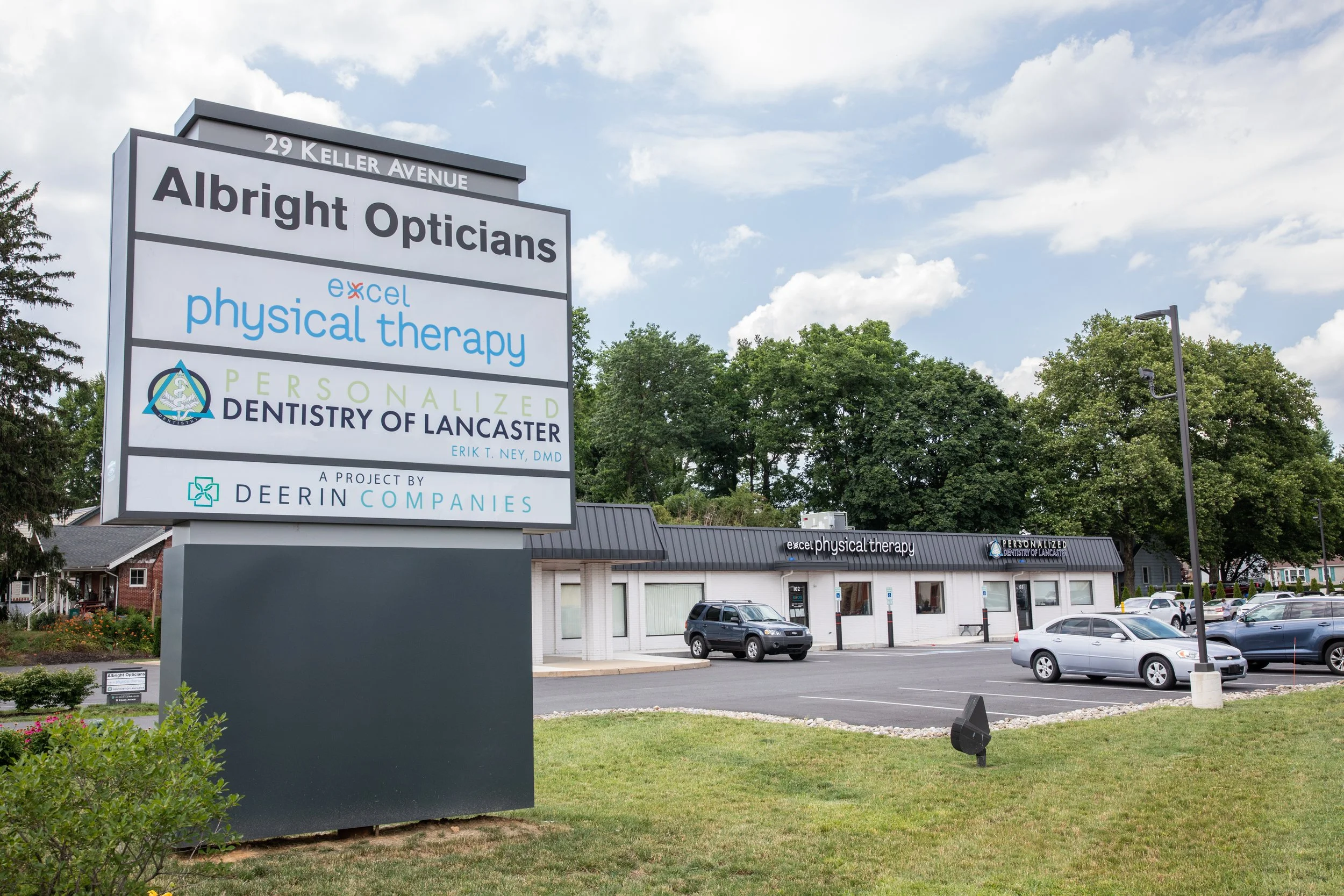A large sign outside a building listing businesses including Albright Opticians, Excel Physical Therapy, and the Personalize Dentistry of Lancaster, on Keller Avenue, with cars parked in front and trees in the background.