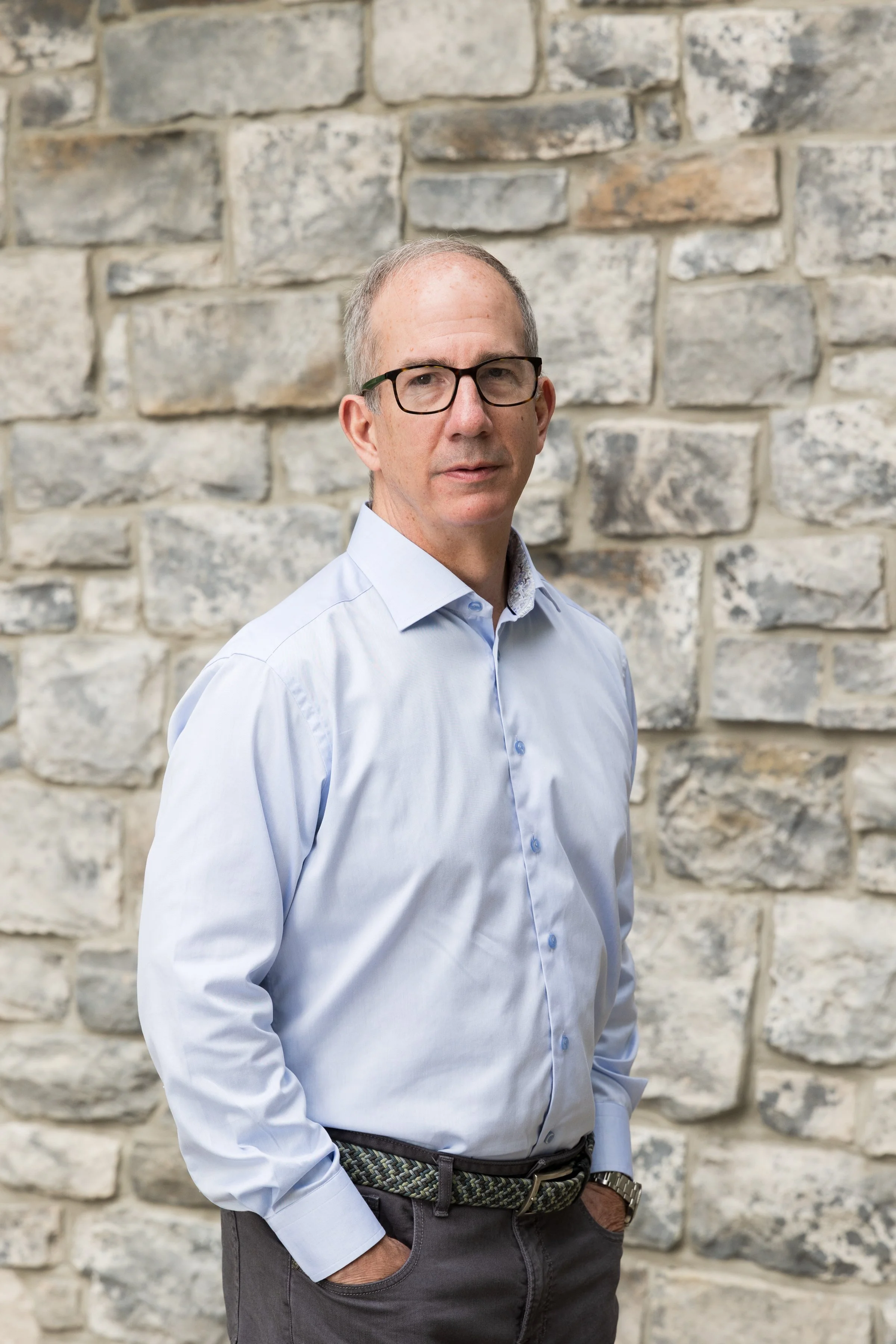 A middle-aged man wearing glasses, a light blue dress shirt, dark pants, and a patterned belt stands with his hands in his pockets in front of a stone wall.