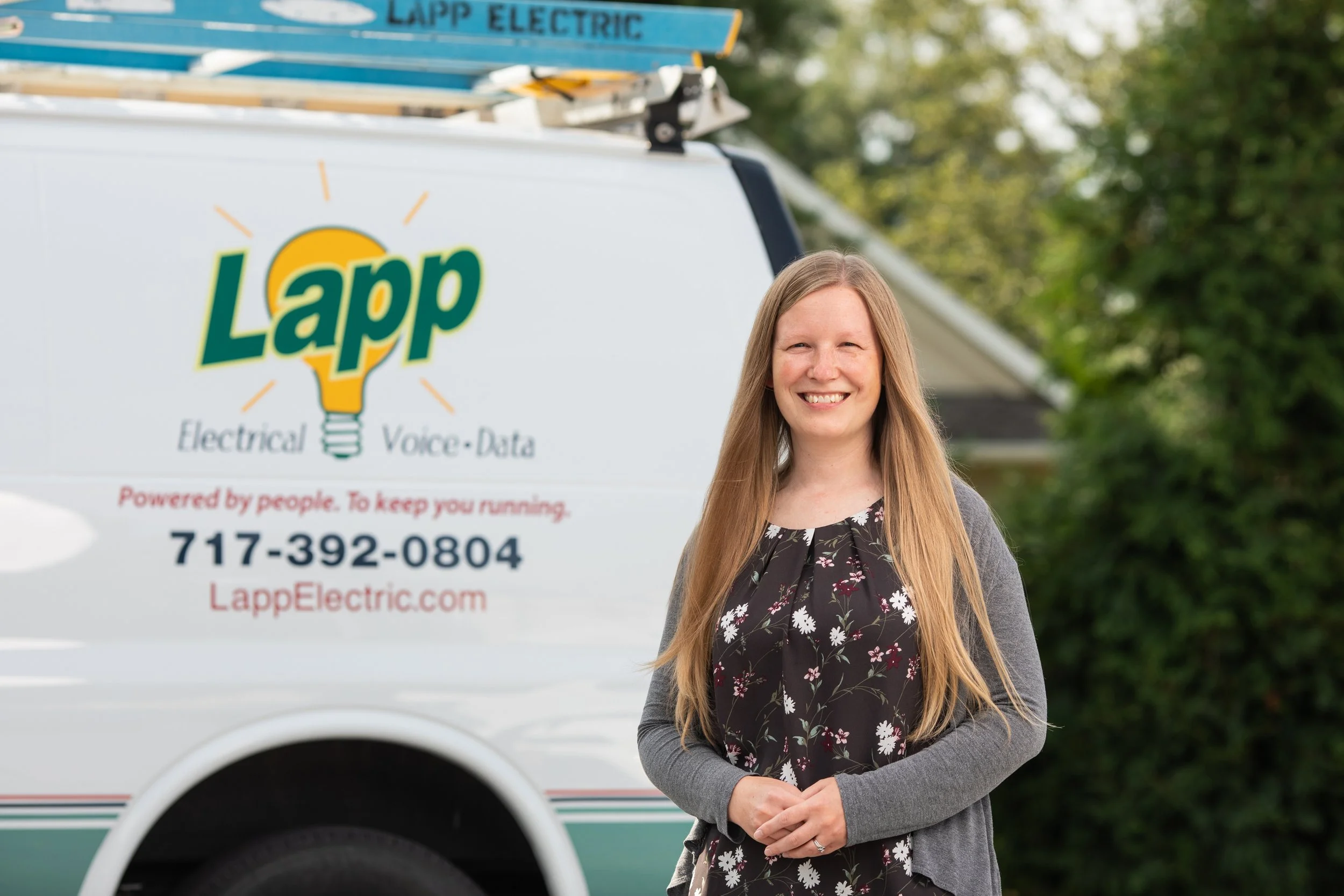 A smiling woman with long light brown hair, wearing a black floral dress and gray cardigan, standing outdoors near a Lapp Electric service van with ladders on top, surrounded by green trees.