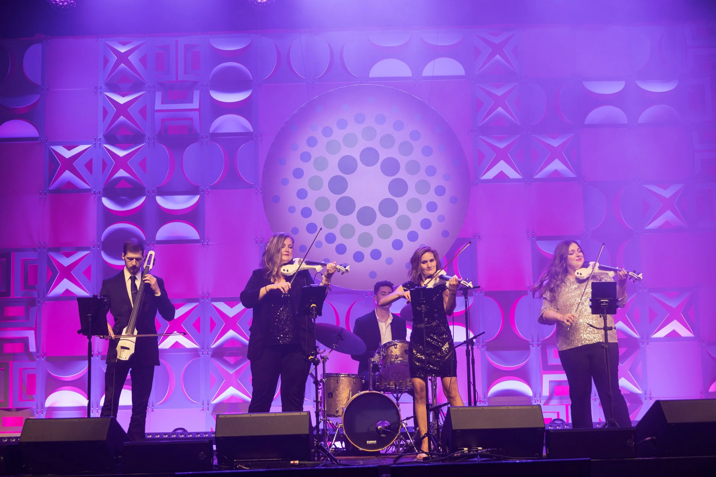 A musical band performing on stage with purple lighting and geometric backdrop. There are five members: a man playing a string instrument, three women playing violins, and a man on drums.