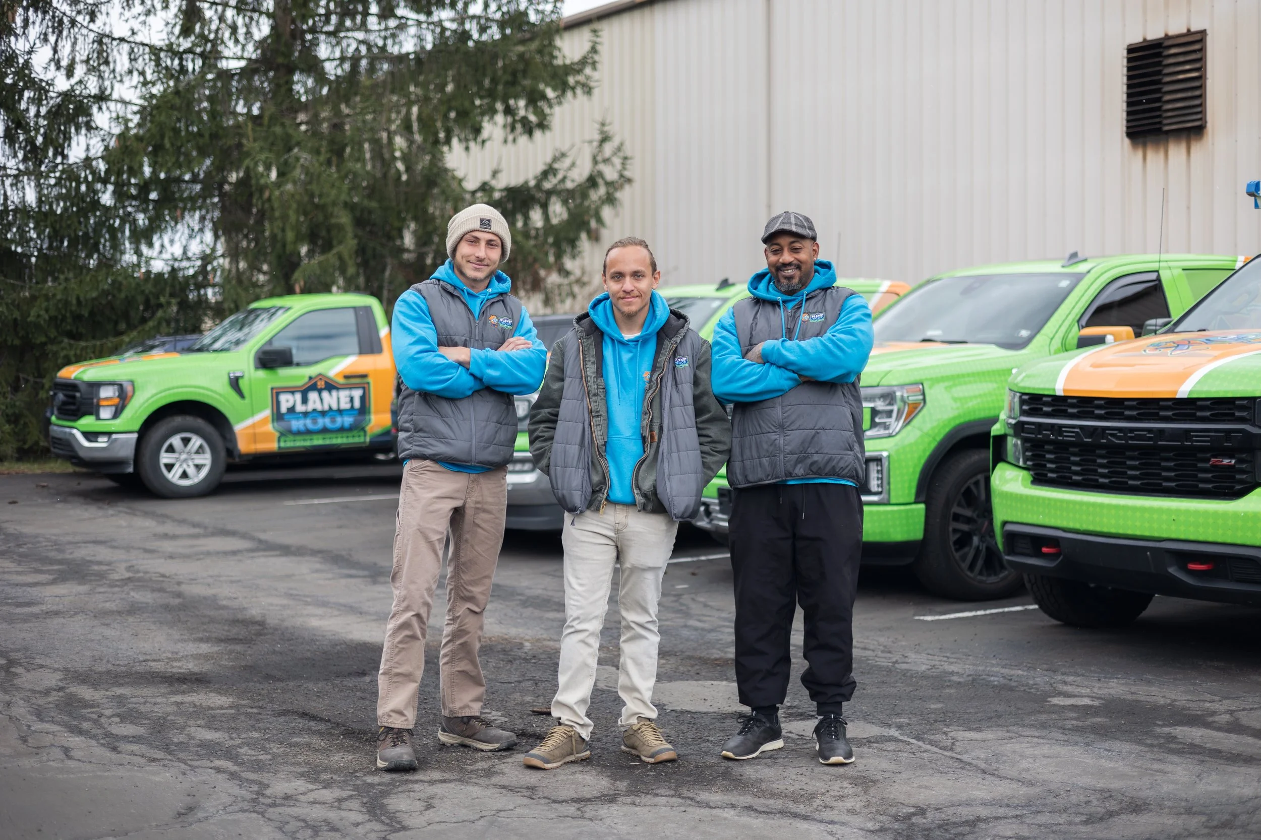 Three men standing in a parking lot with green vehicles behind them, wearing blue hoodies and vests with the 'Planet Roof' logo.