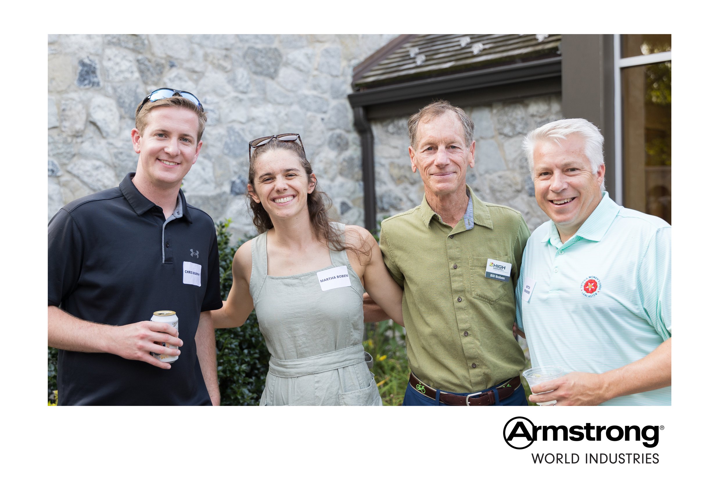 Four people posing outdoors at a social event, with a stone building background, smiling and holding drinks, wearing name tags, with 'Armstrong World Industries' logo at the bottom.