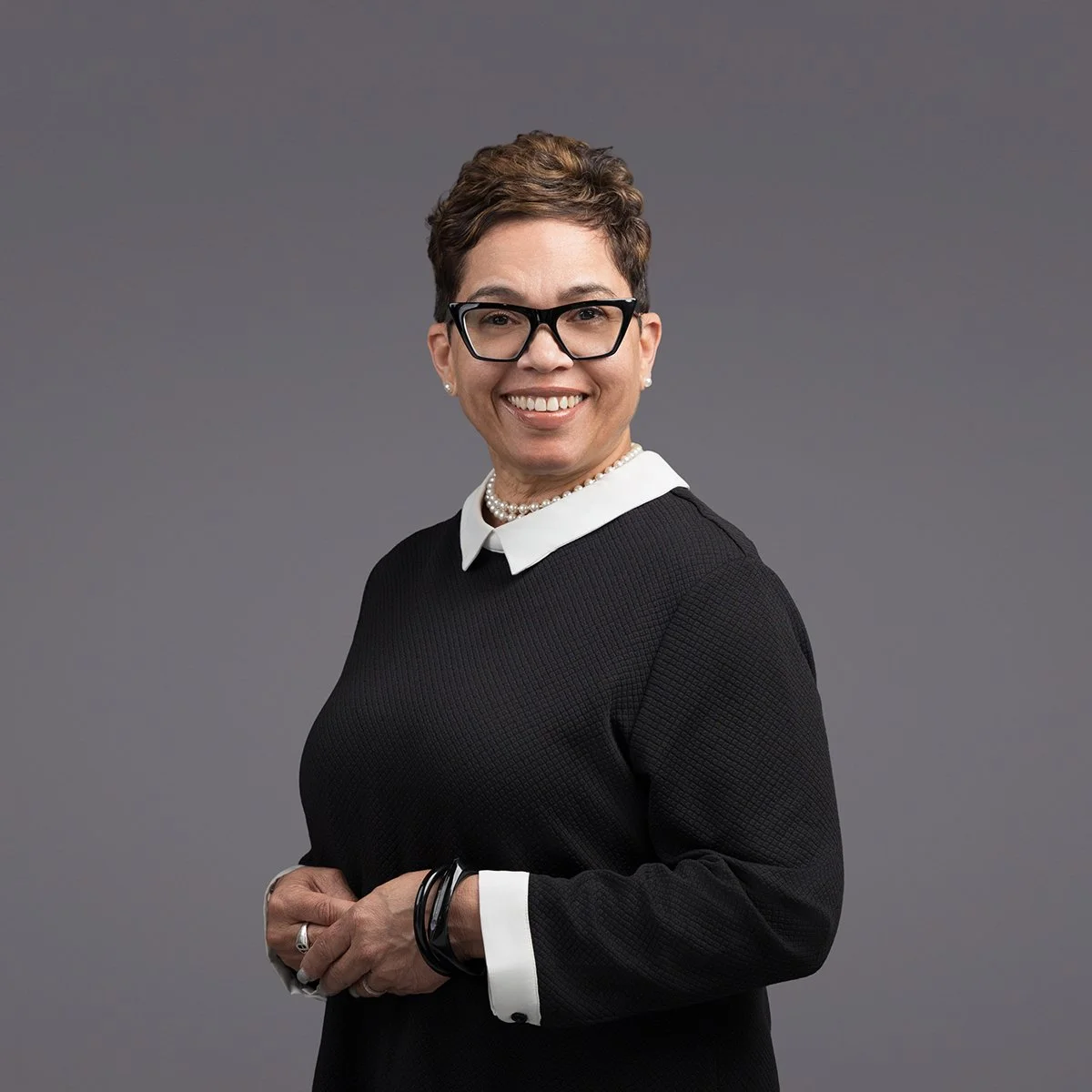 A woman with short brown hair, black glasses, wearing a black sweater with a white collar, necklaces, and bracelets, smiling against a gray background.