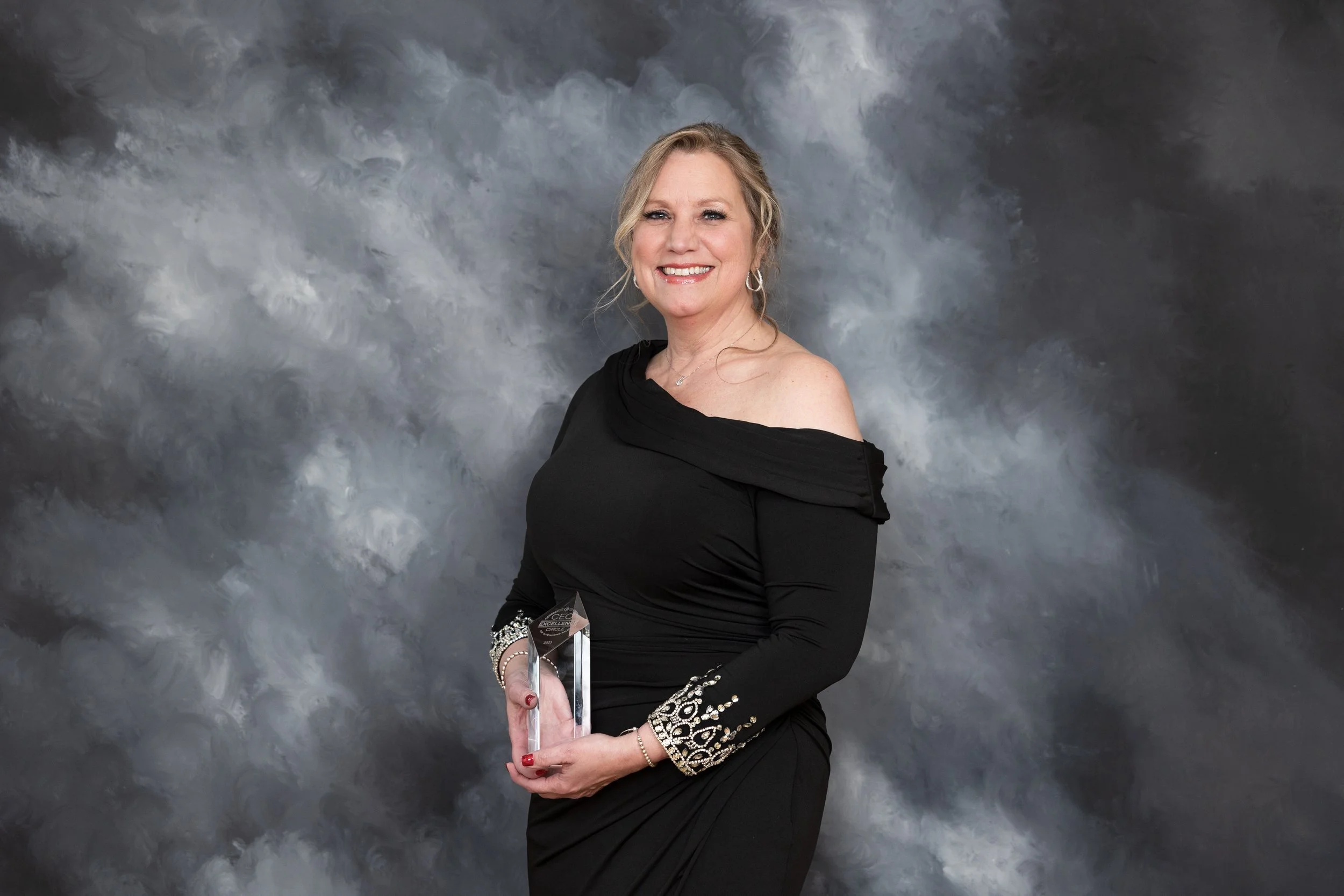A woman in a black off-shoulder dress holding an award, smiling, with a cloudy gray background.