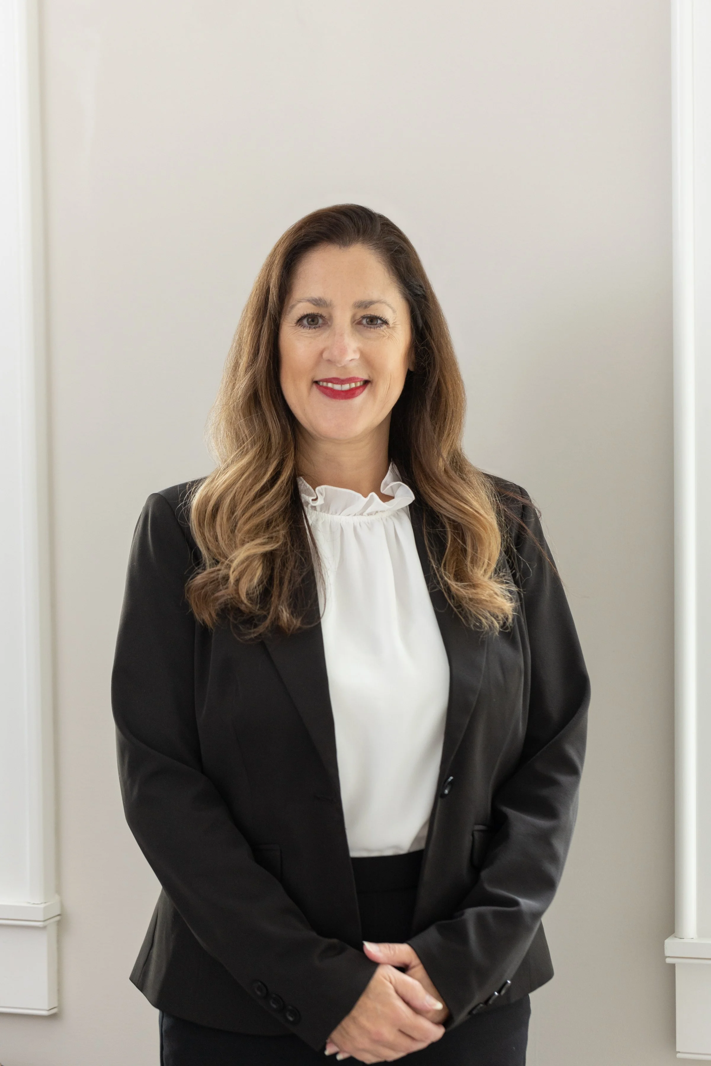 A professional woman with long, wavy brown hair, wearing a black blazer over a white blouse, standing indoors with a neutral background.