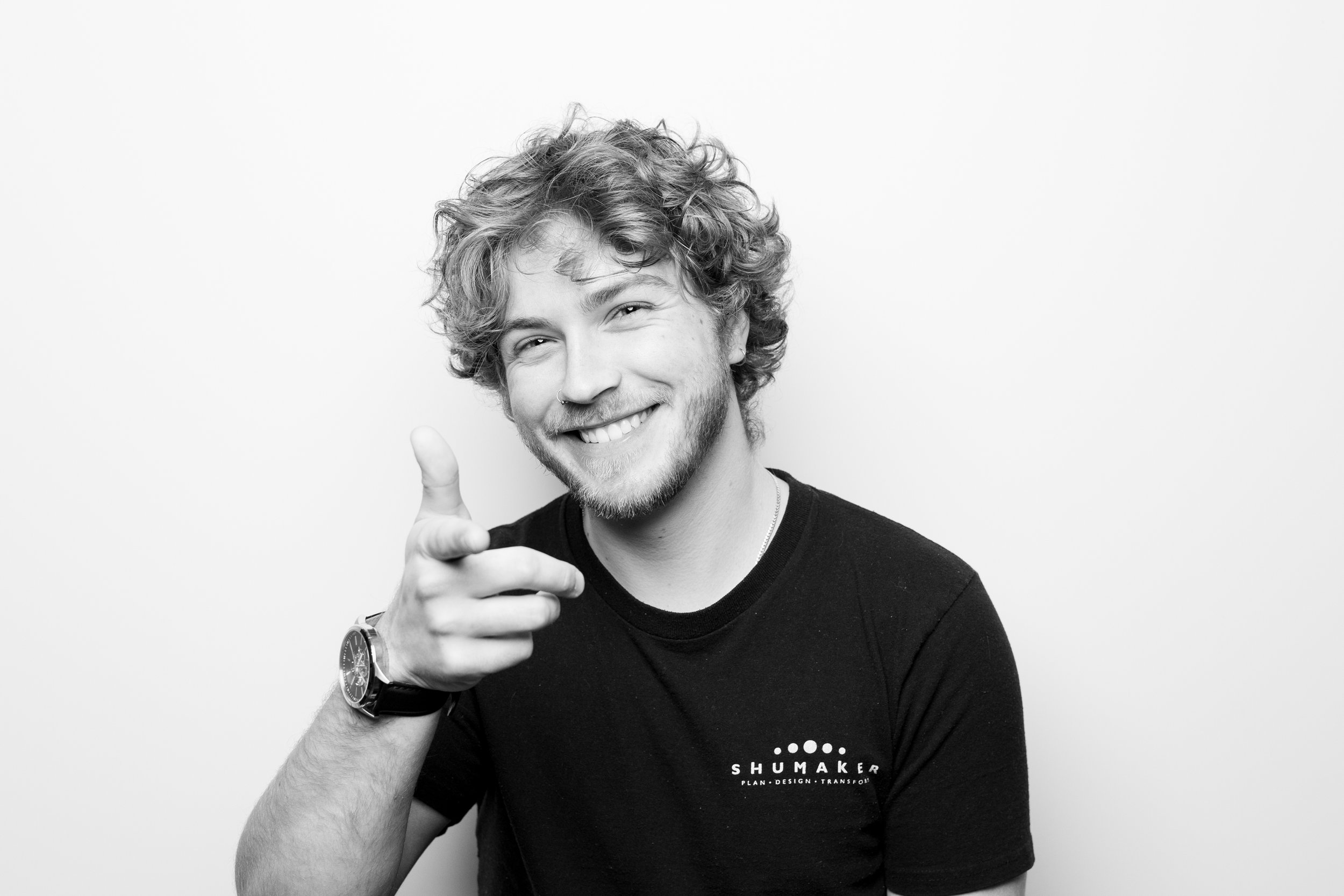 Black and white photo of a smiling young man with curly hair, pointing towards the camera, wearing a black t-shirt with the word "SHUMAKER" and smaller text underneath, a watch on his left wrist, and a nose ring.