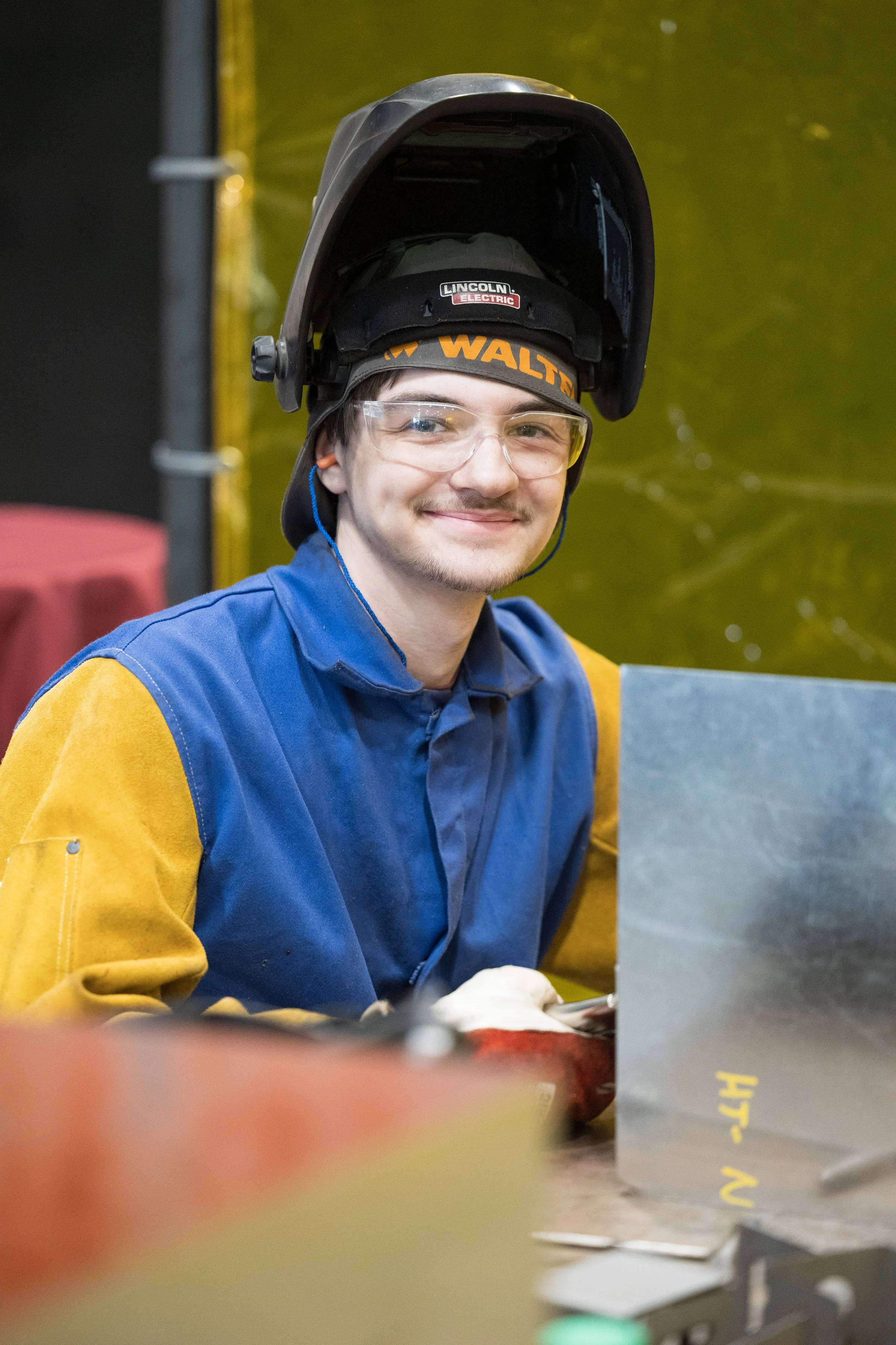 A young man in protective welding gear, including a helmet, goggles, and a work coat, smiling at the camera in an industrial or workshop setting.