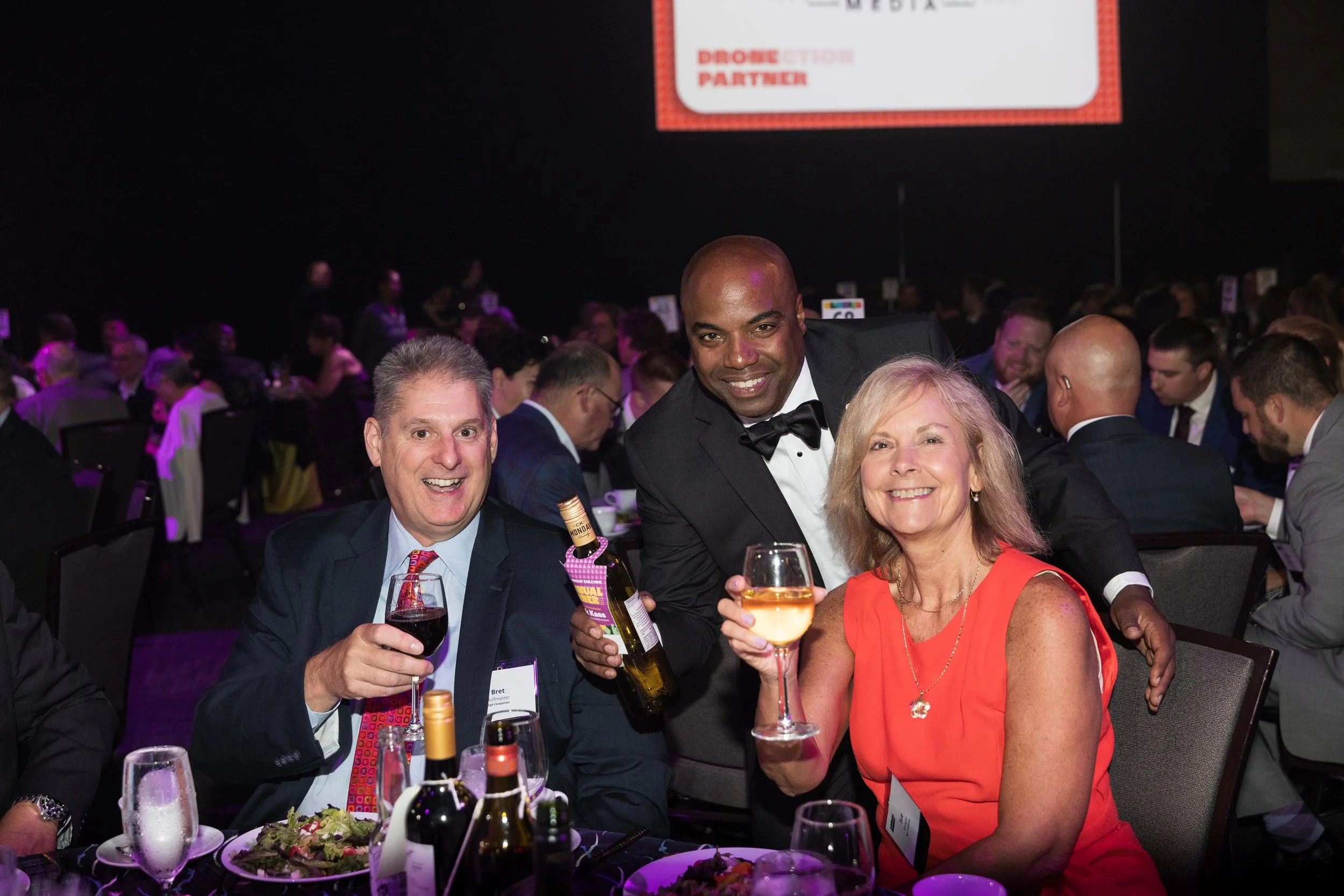 Three people celebrating at a banquet, smiling and holding drinks, with a crowd in the background.