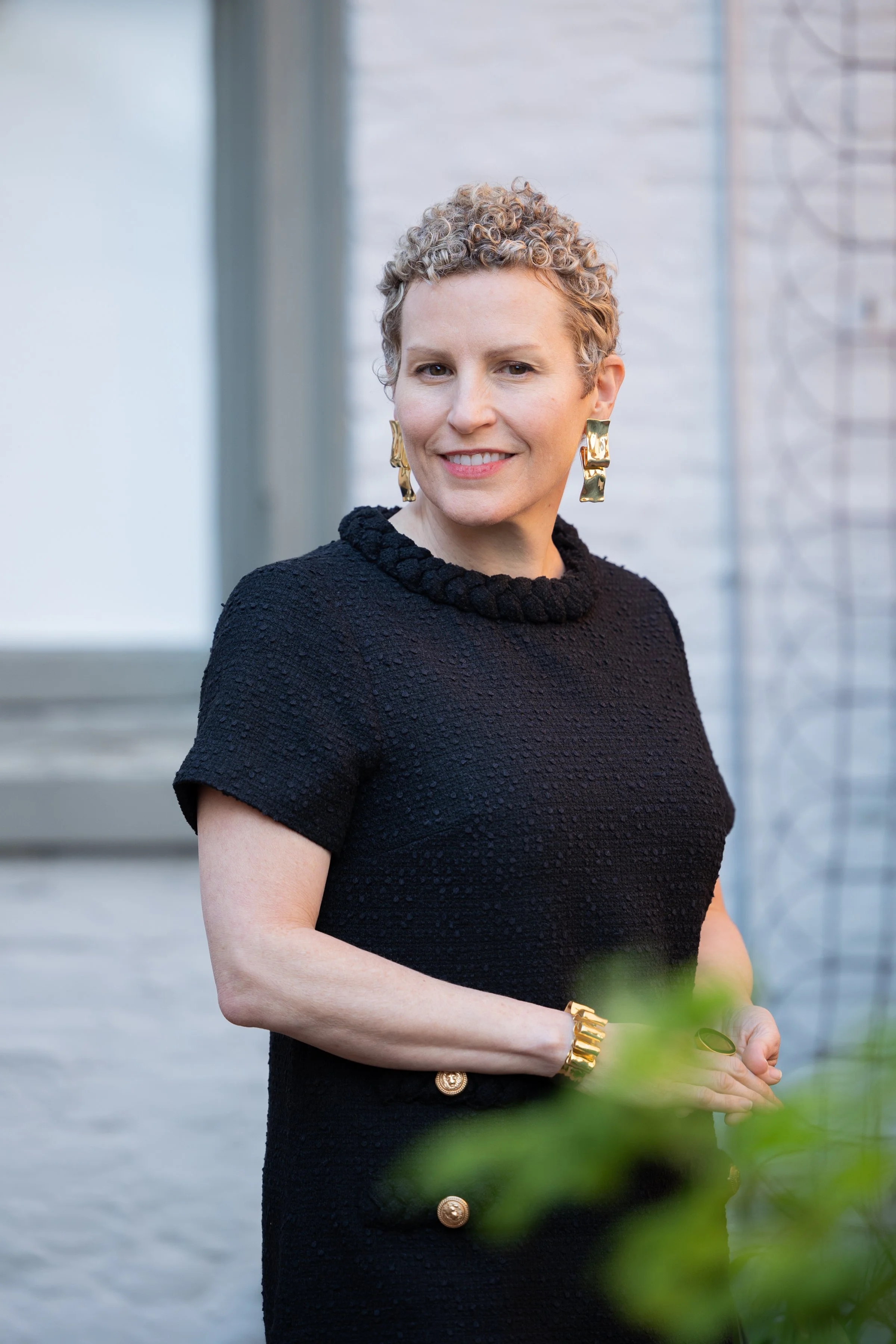 A woman with short curly blonde hair, wearing a black textured dress with gold buttons, large gold earrings, and a gold bracelet. She is smiling and standing outdoors near a white wall and window.