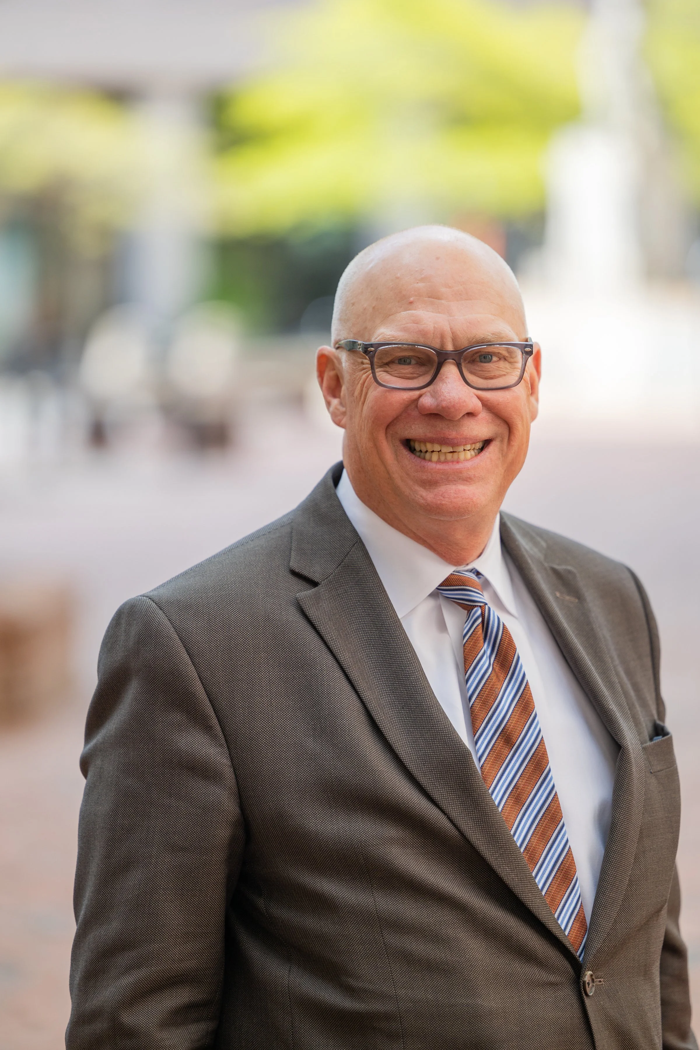 A smiling middle-aged man wearing glasses, a white shirt, a brown suit, and a striped tie, standing outdoors with a blurred background of trees and buildings.