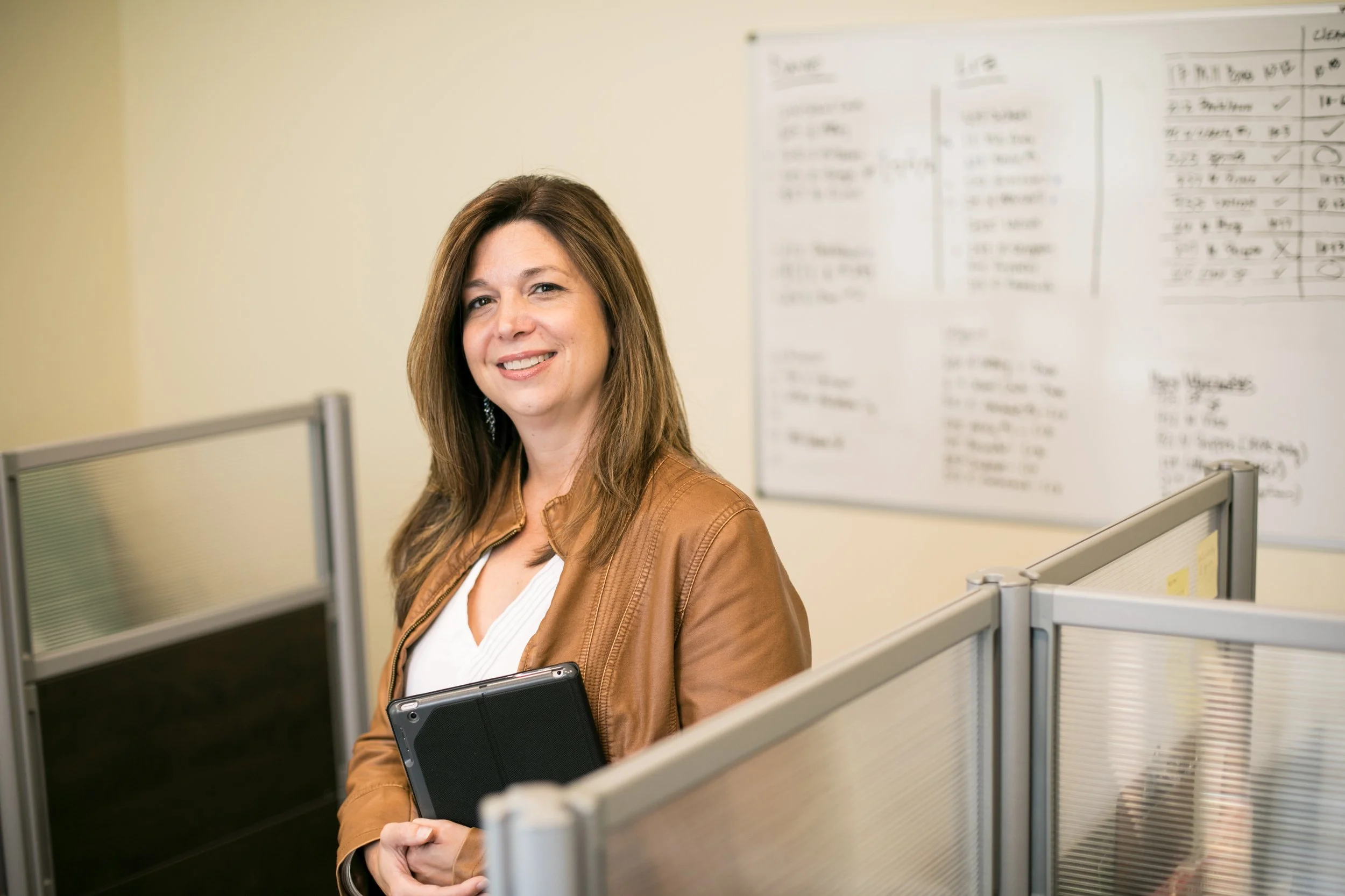 A woman smiling and holding a tablet in an office cubicle, with a whiteboard filled with notes and a schedule behind her.