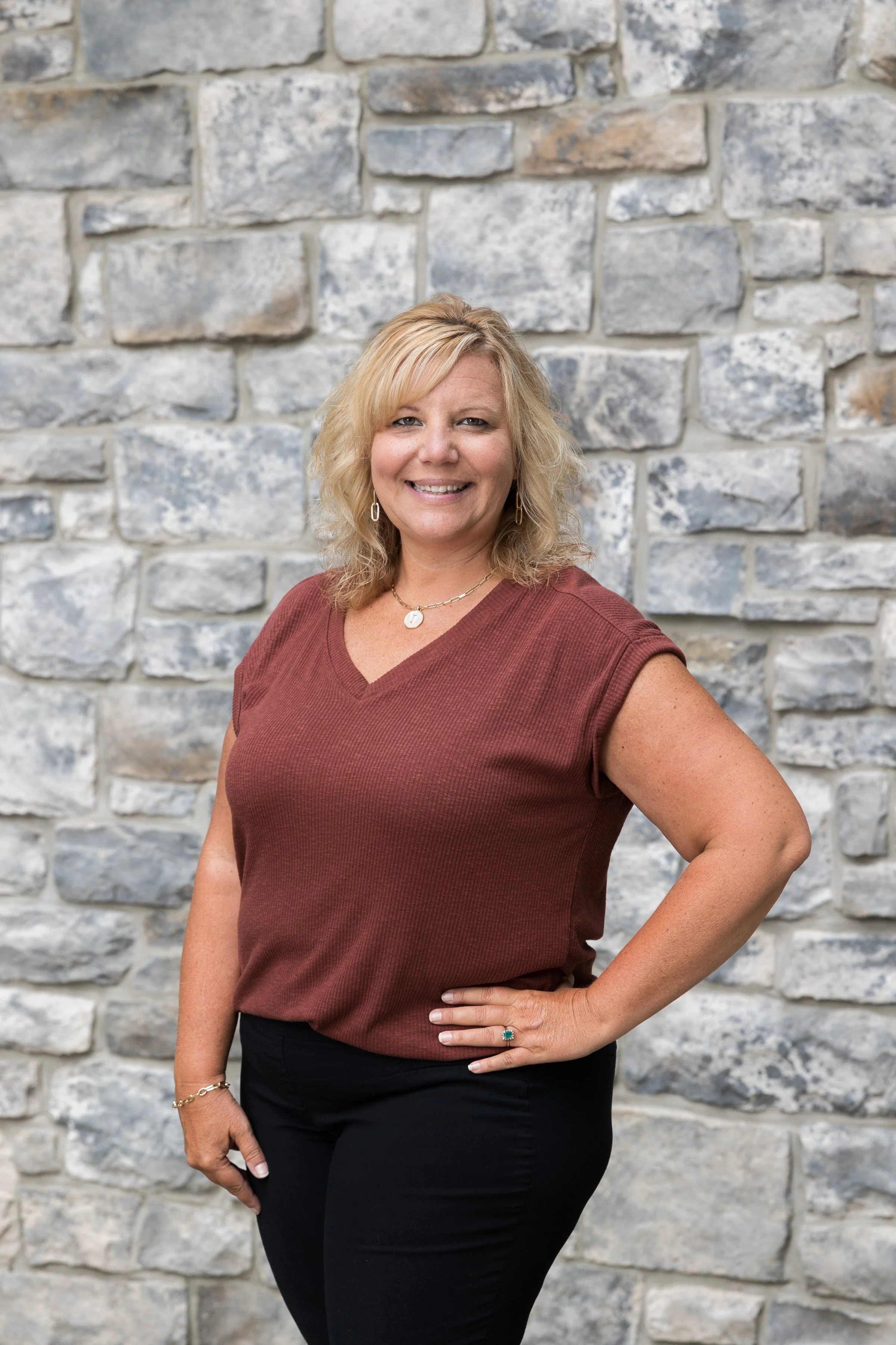 A woman with blonde, curly hair wearing a maroon short-sleeve top and black pants standing against a stone wall, smiling with her hand on her hip.