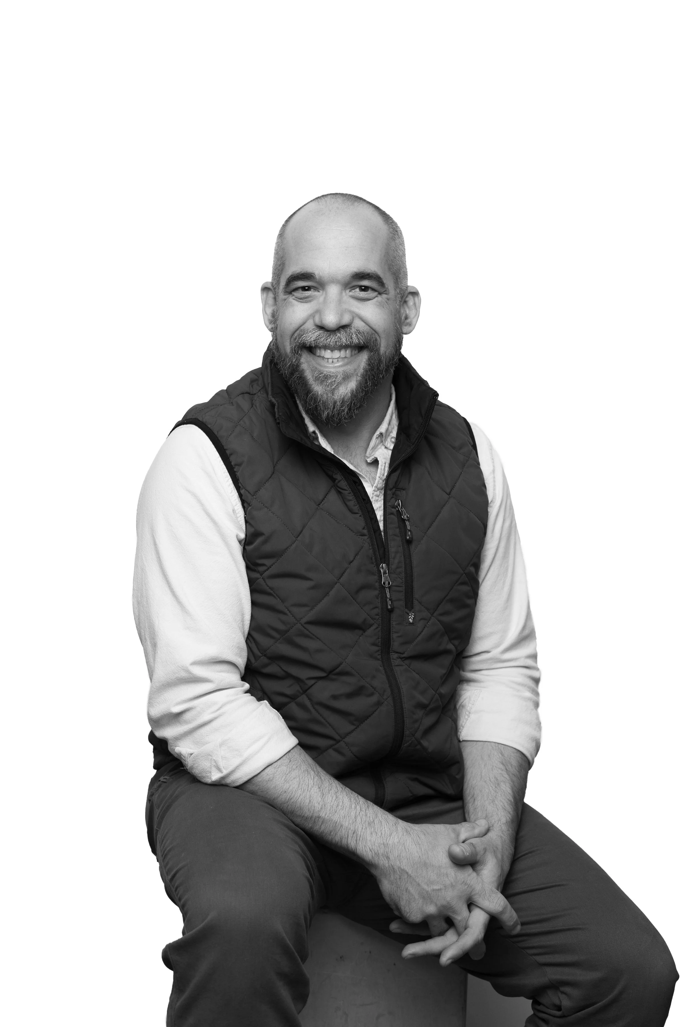Black and white portrait of a man with a beard, smiling, wearing a vest over a long-sleeve shirt, sitting on a stool isolated on a white background.