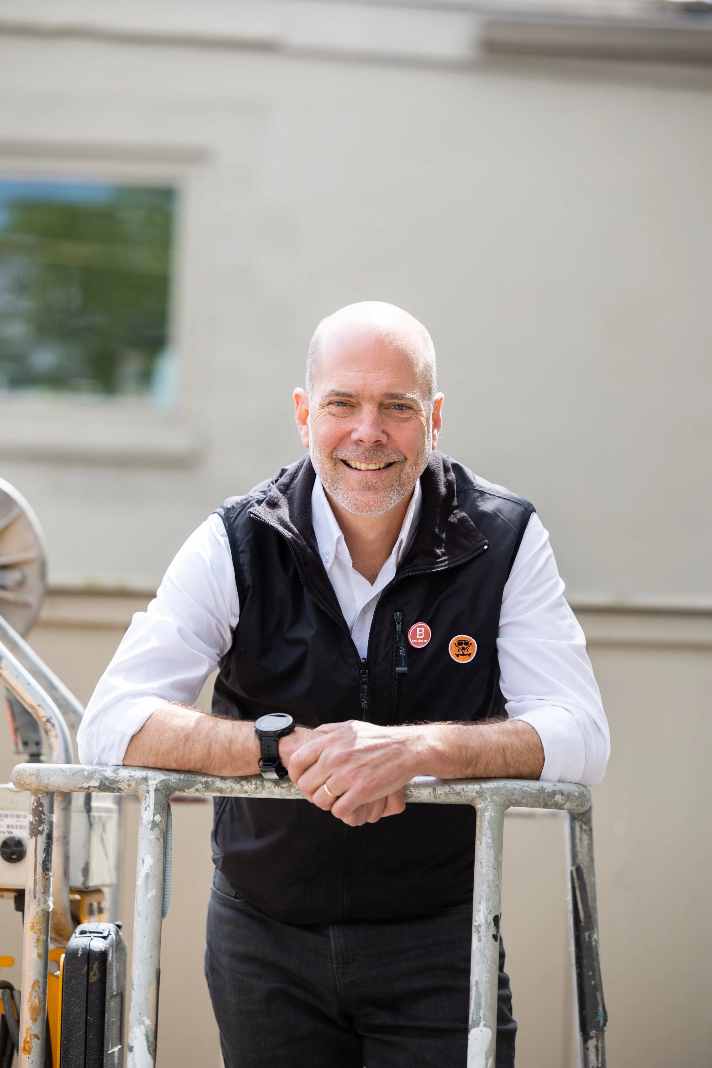 A middle-aged man with a bald head and a beard, smiling, wearing a white shirt and a black vest, leaning on a metal scaffold outdoors.