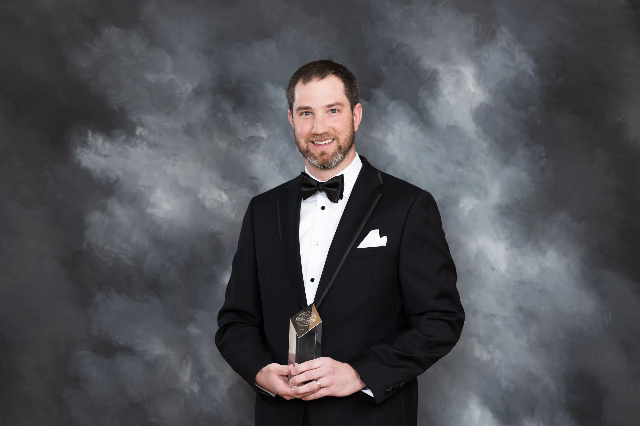 A man in a black tuxedo with a bow tie holding an award, standing against a gray cloudy background.