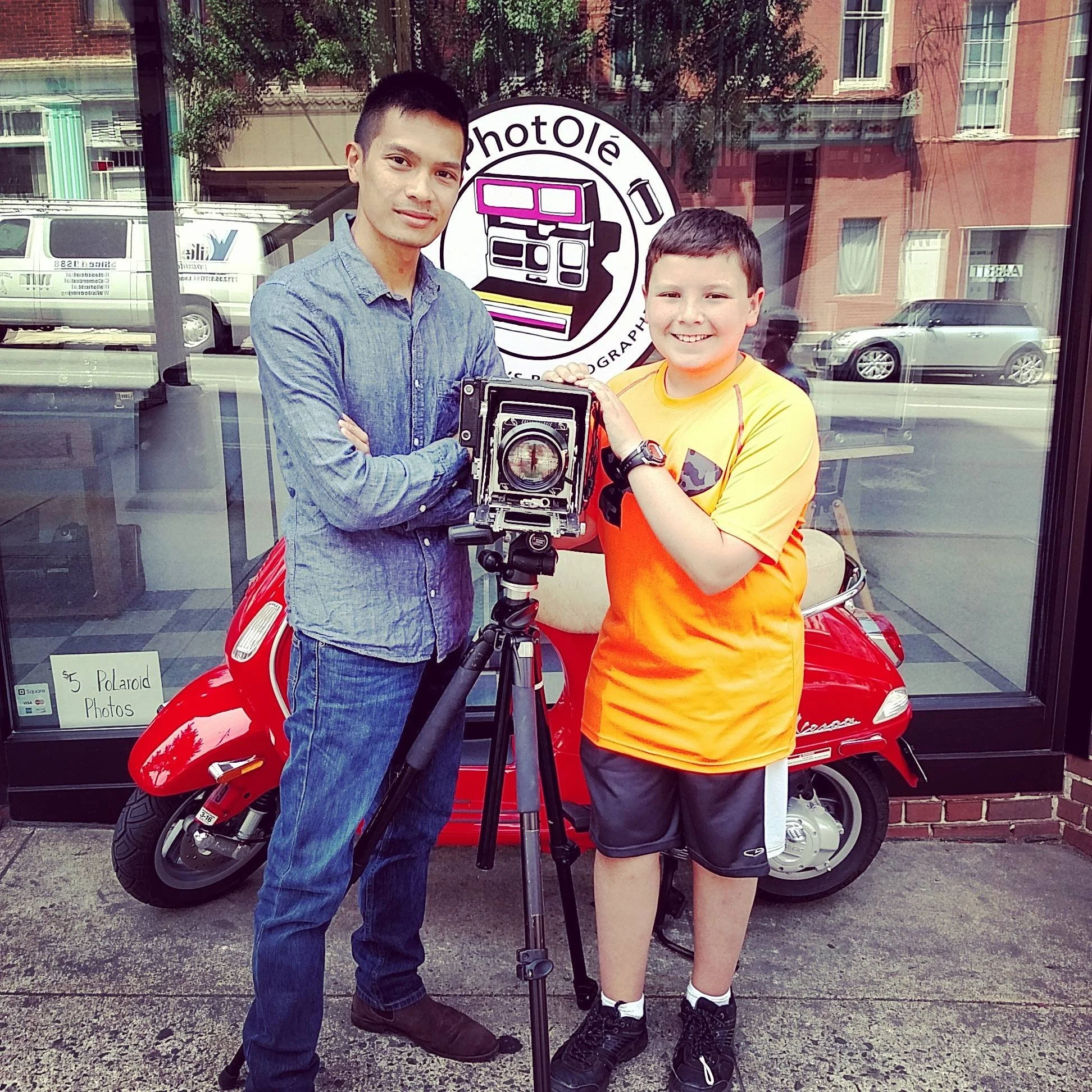 Two boys standing outside a photo studio, one holding a vintage camera mounted on a tripod, with a red Vespa scooter and studio window with sign in the background.