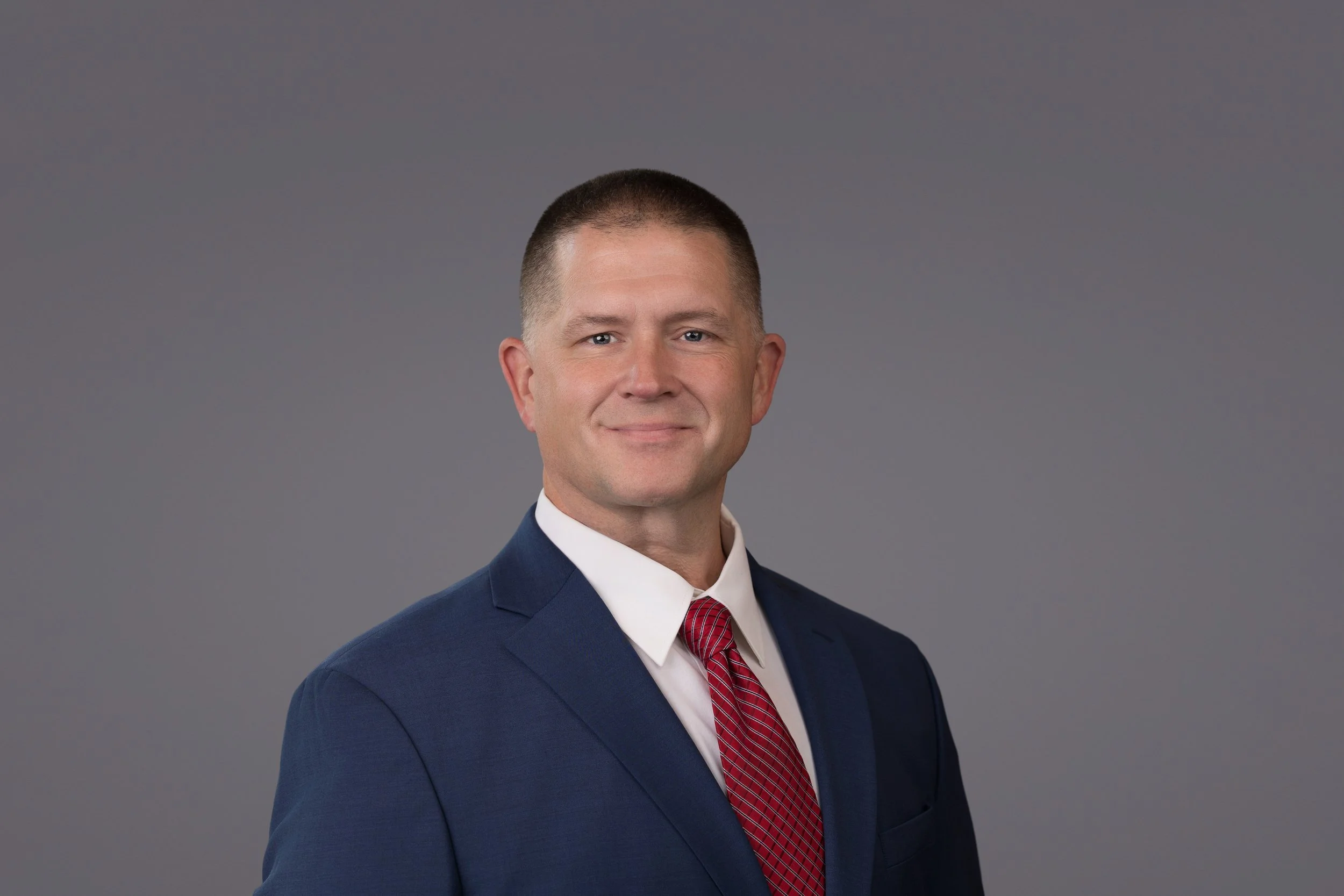 Professional headshot of a man in a blue suit, white shirt, and red tie, against a gray background.