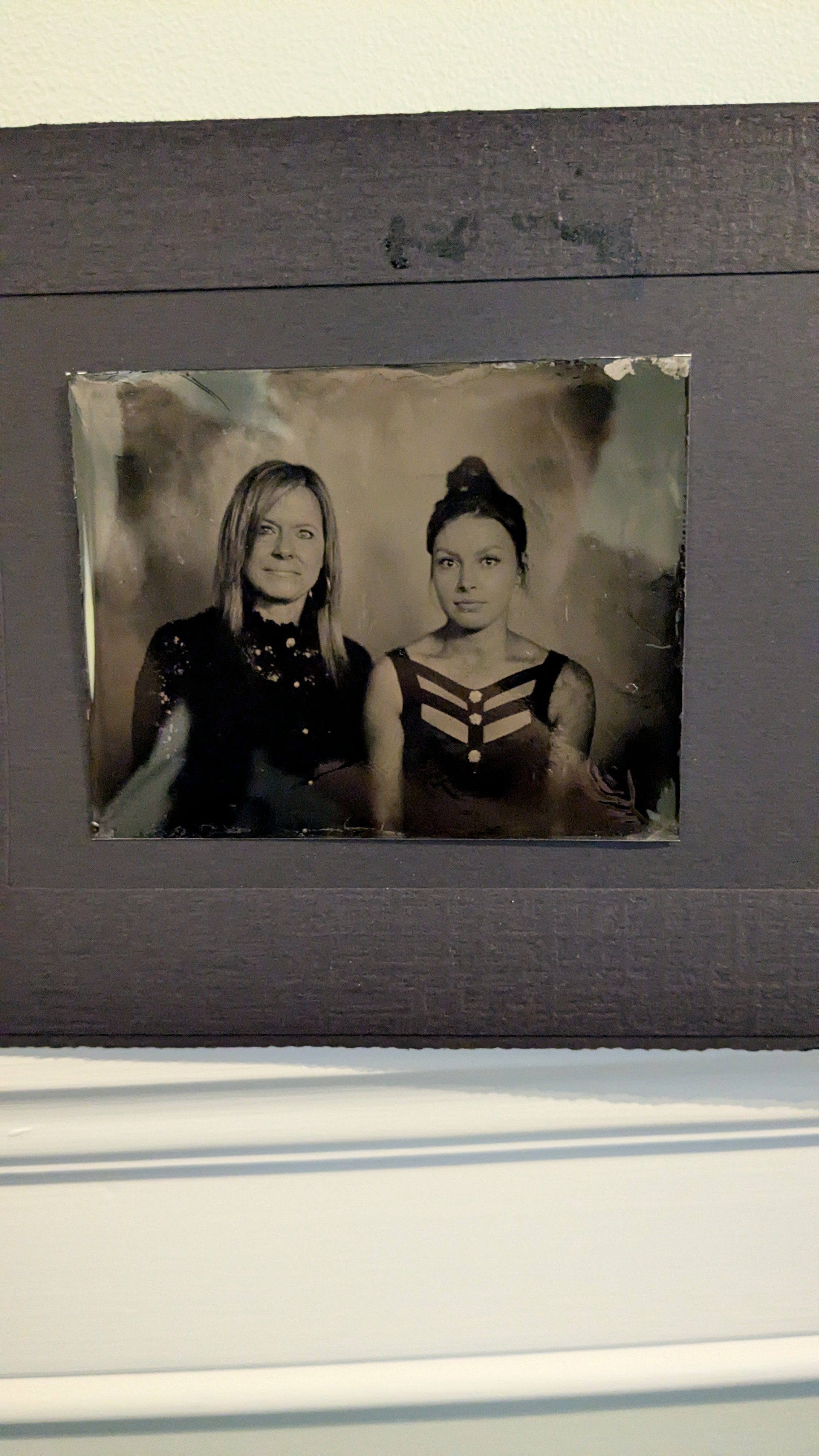 Black and white framed photograph of two women, one with shoulder-length hair wearing a dark lace dress, and the other with hair in a bun wearing a patterned dress with cutouts, against a cloudy background.
