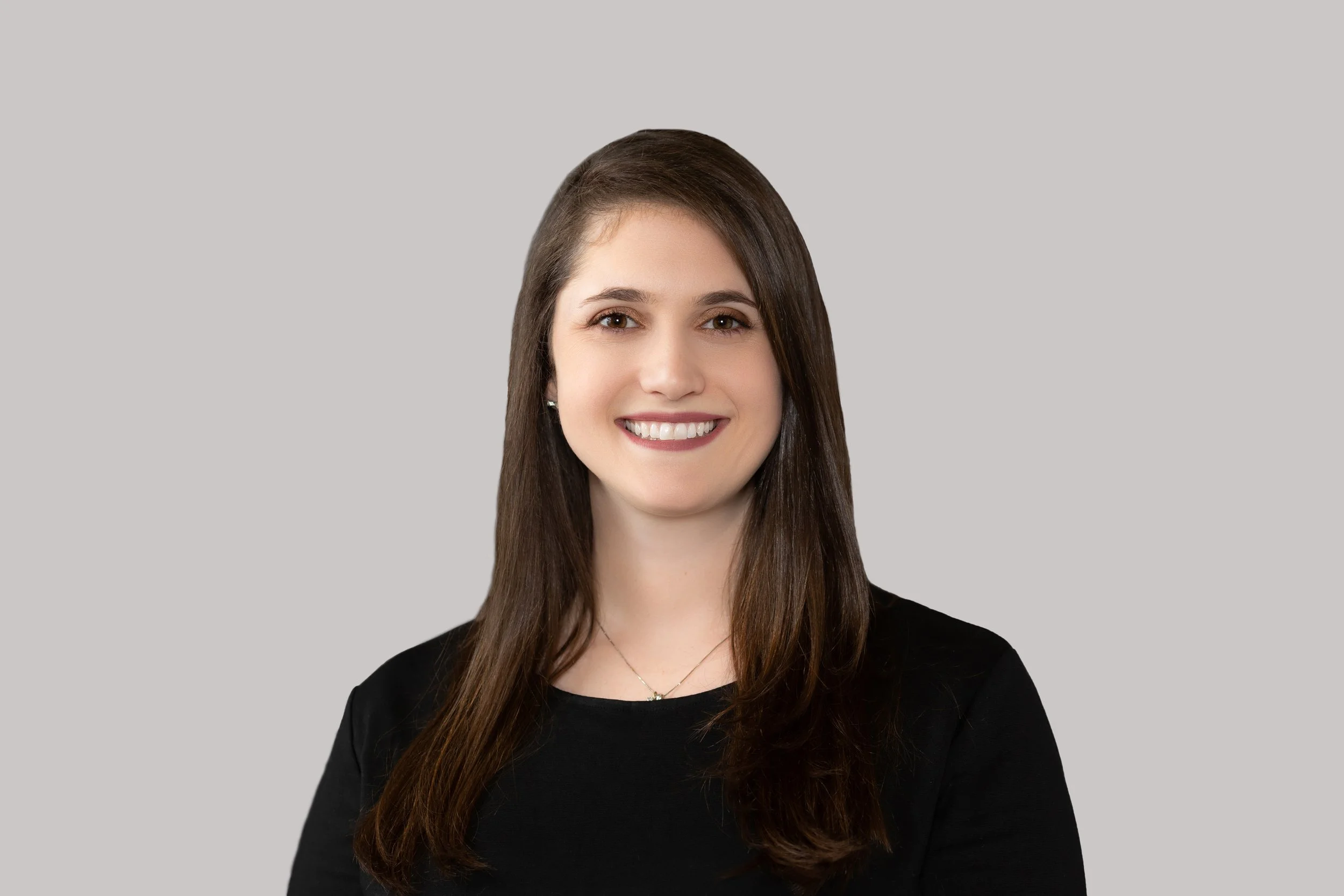 A smiling woman with long brown hair wearing a black top and a small necklace, against a plain gray background.