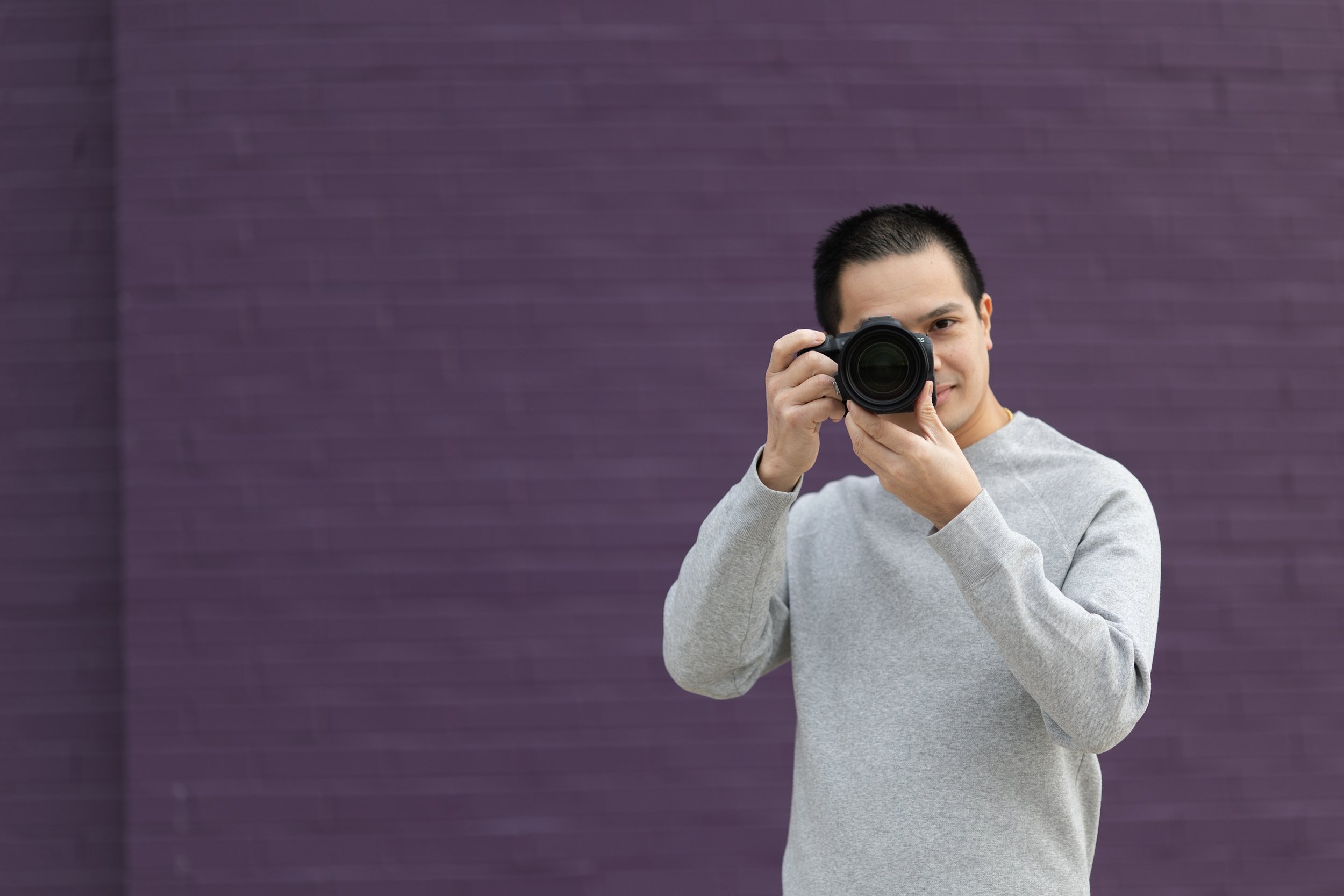 A man in a gray sweater holding a camera, taking a picture, standing in front of a purple wall.