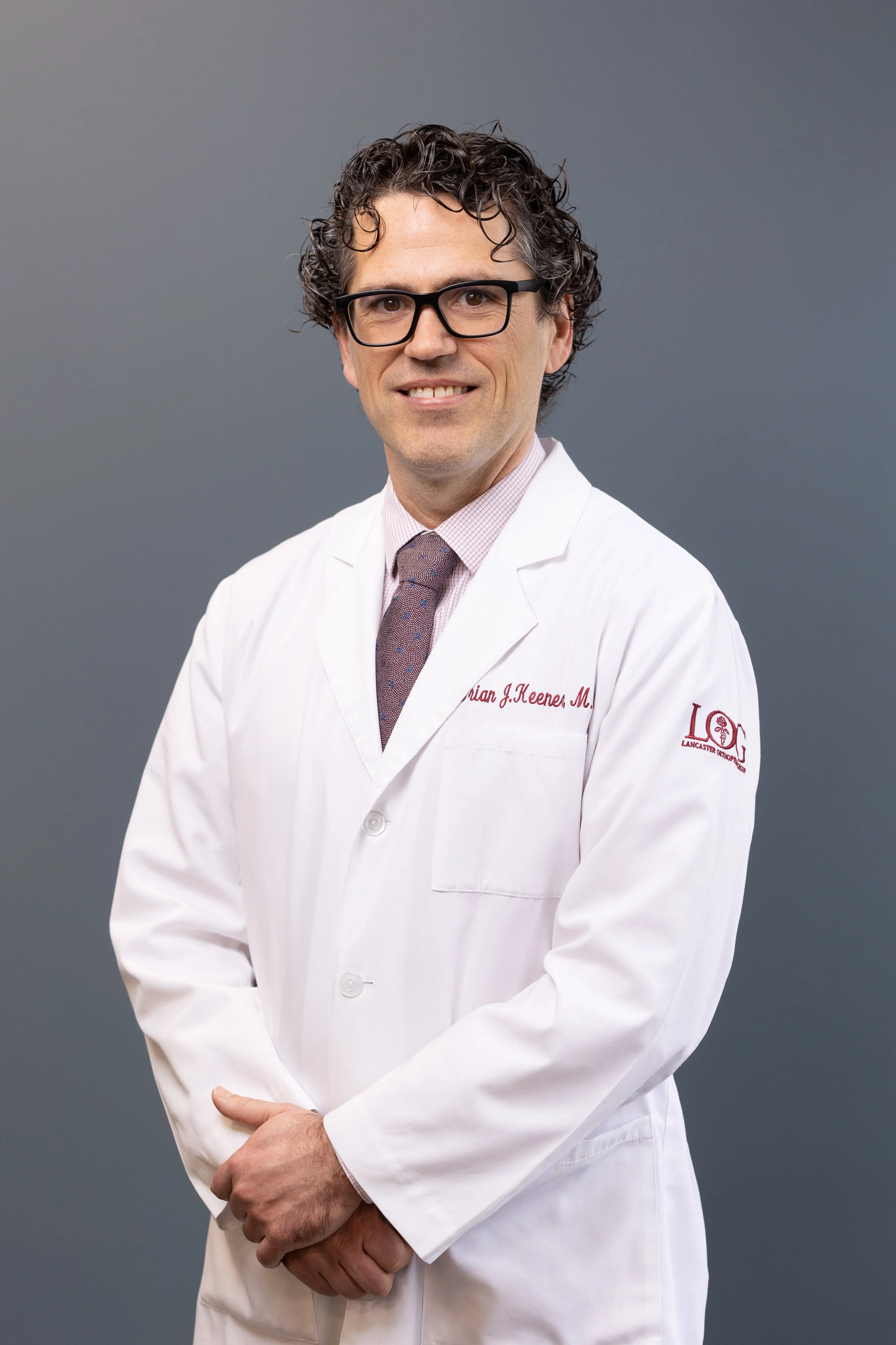 A man with curly dark hair, glasses, wearing a white lab coat and a patterned tie, smiling against a plain grey background.