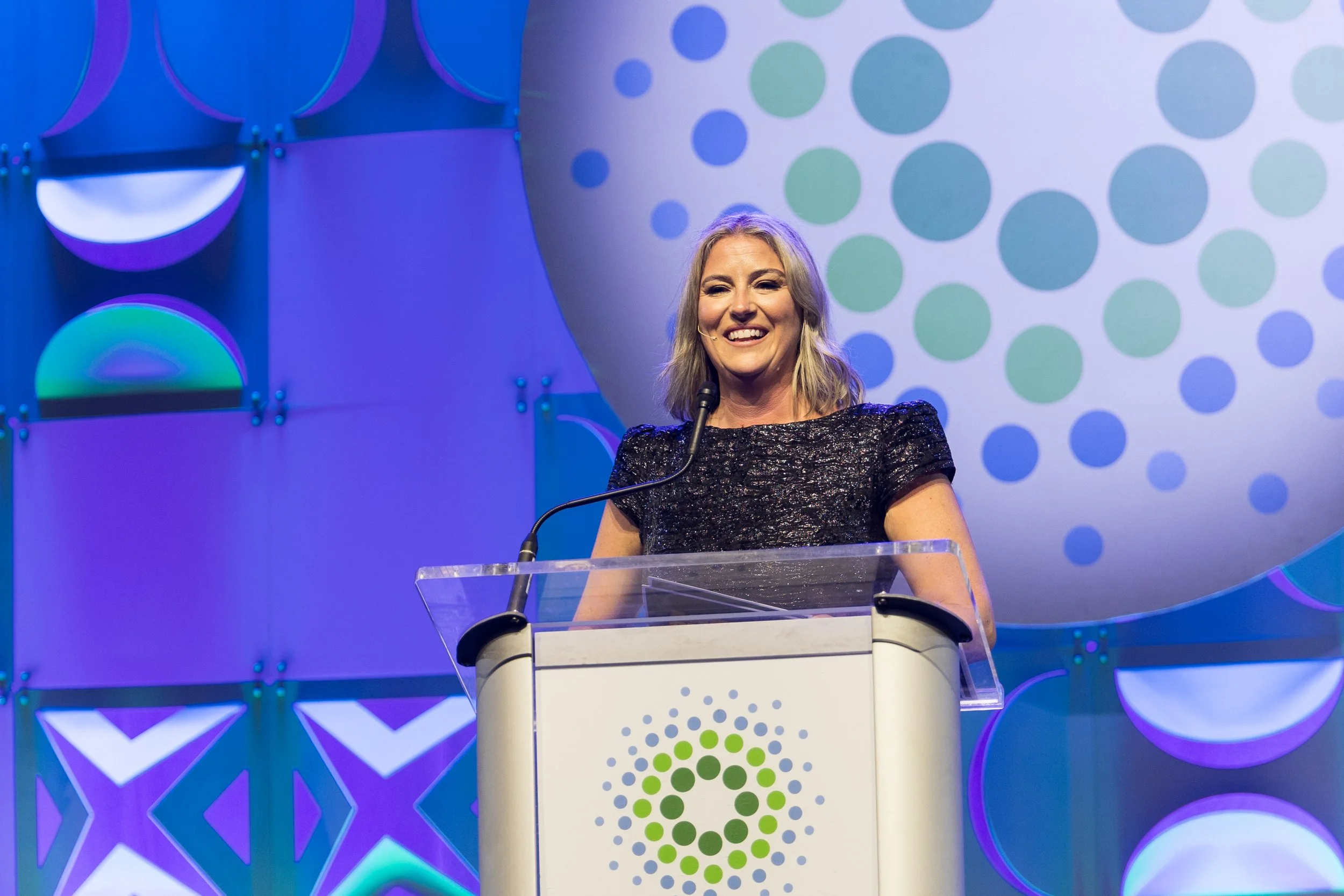 A woman with blonde hair stands at a podium, smiling, at an event with a colorful, abstract backdrop featuring circles and geometric shapes.