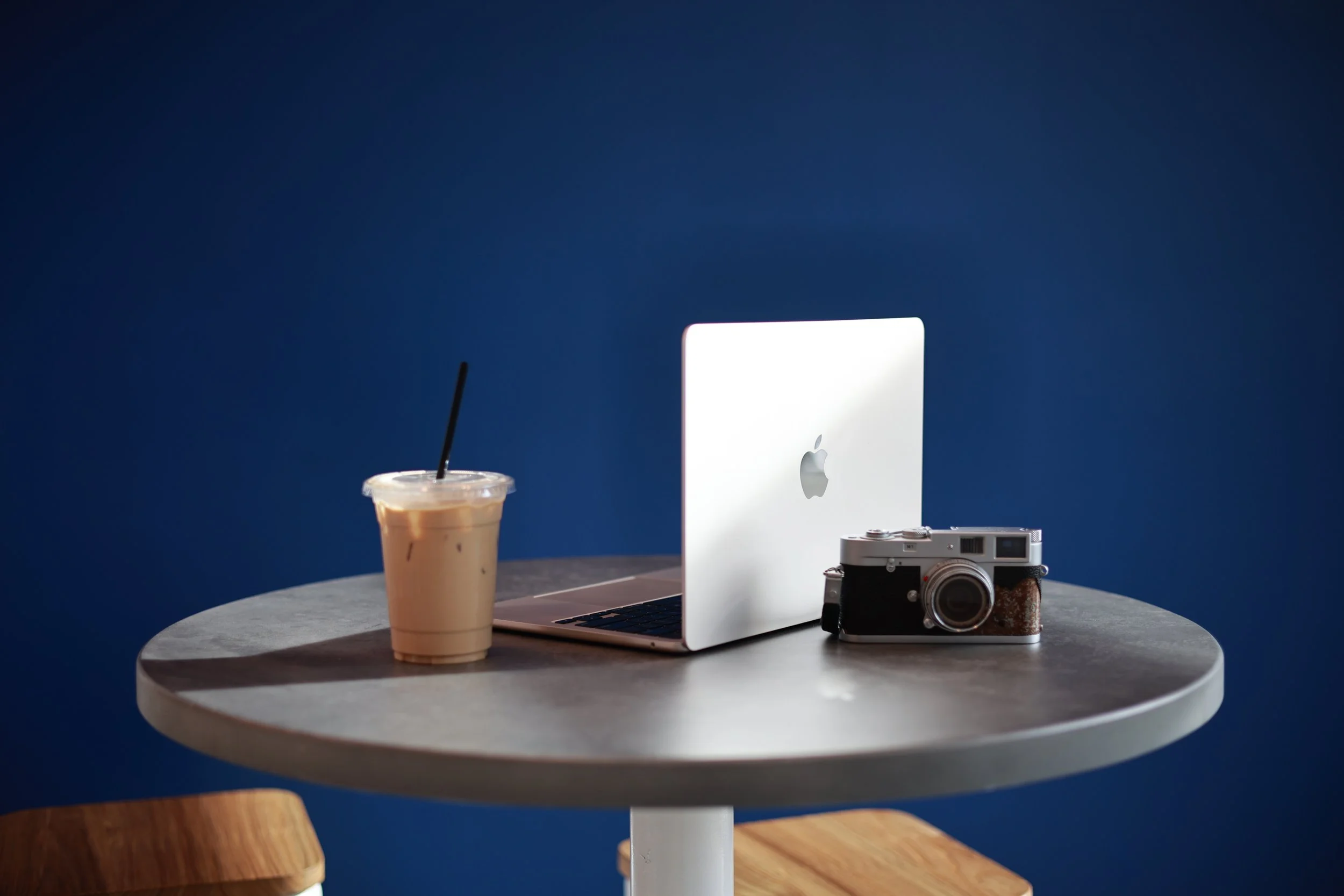 A round table with a iced coffee, a silver Apple MacBook, and a vintage camera against a dark blue background.