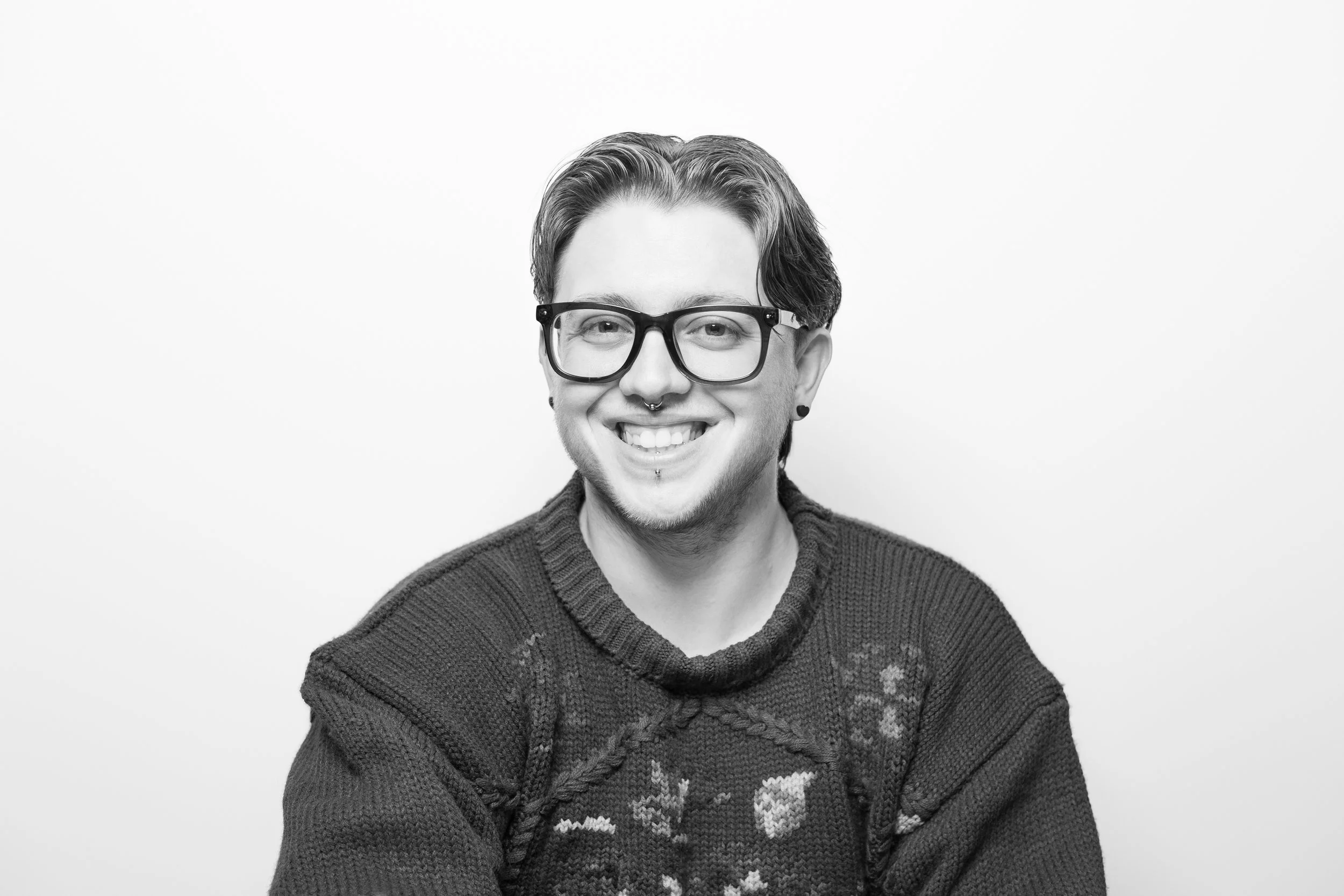 Black and white portrait of a smiling young man with glasses, piercings, and a patterned sweater, against a plain white background.