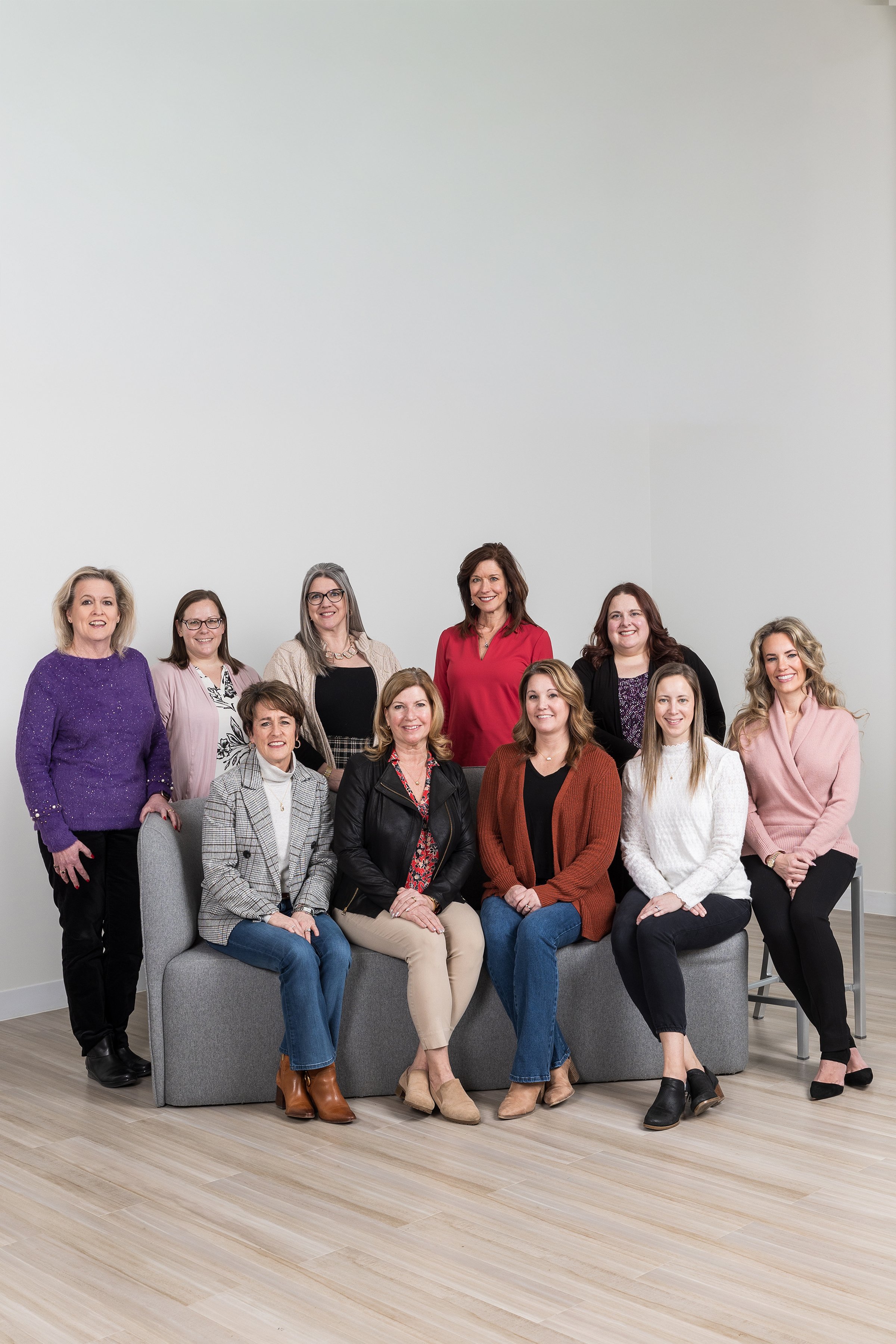 Group portrait of ten women posing together in an indoor setting, some seated on a gray couch and others standing behind it, with a plain white wall in the background.