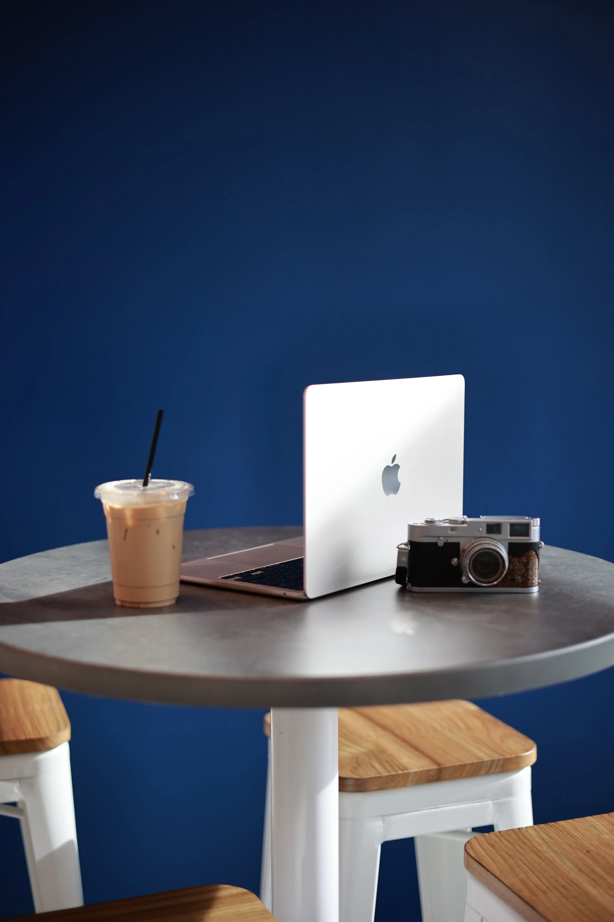 A round table with a MacBook, a vintage camera, and iced coffee on it, against a blue wall.