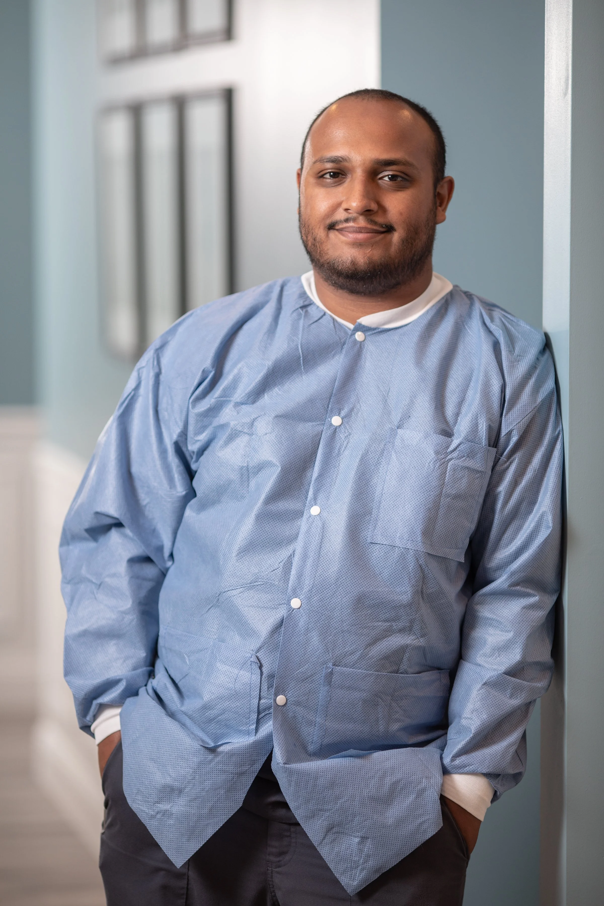 A man with short hair and a beard leaning against a wall in a medical setting, wearing a blue medical gown and smiling.