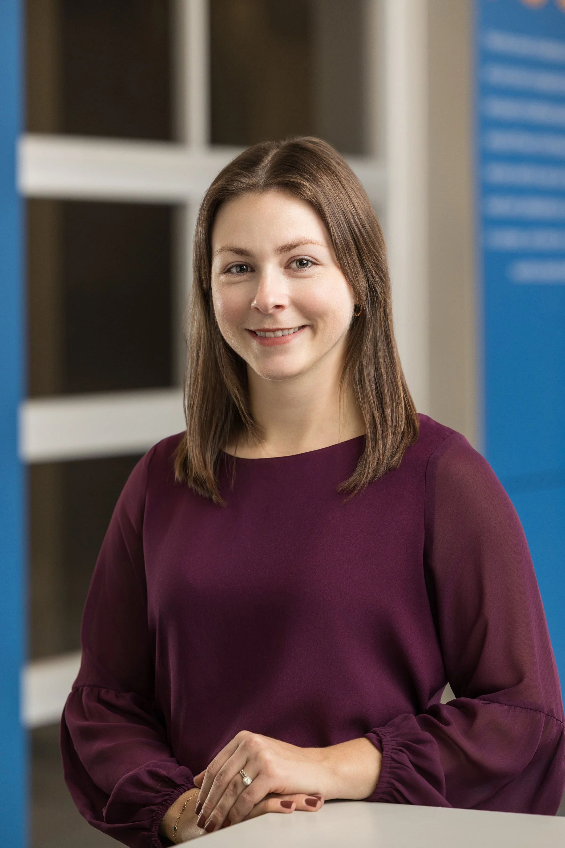 A young woman with shoulder-length brown hair, smiling, sitting at a table in an indoor setting, wearing a maroon blouse with sheer sleeves.