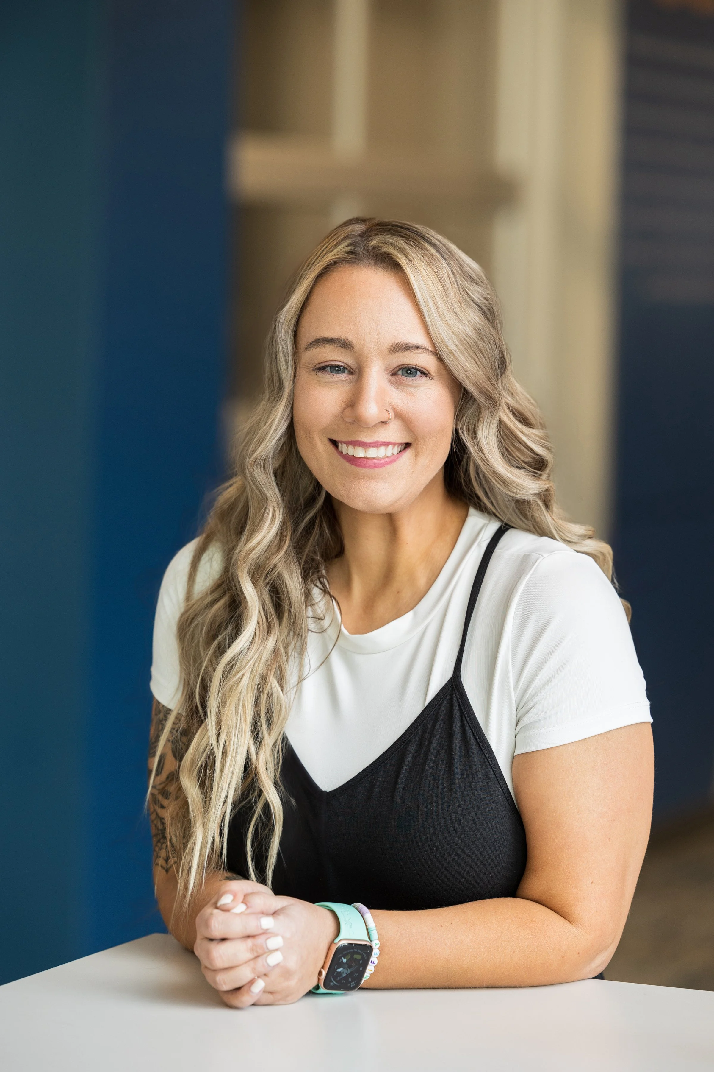 A young woman with long blonde hair, smiling, wearing a white t-shirt under a black spaghetti strap top, sitting at a white table with her hands clasped, wearing a smartwatch and bracelets, in a modern indoor setting.